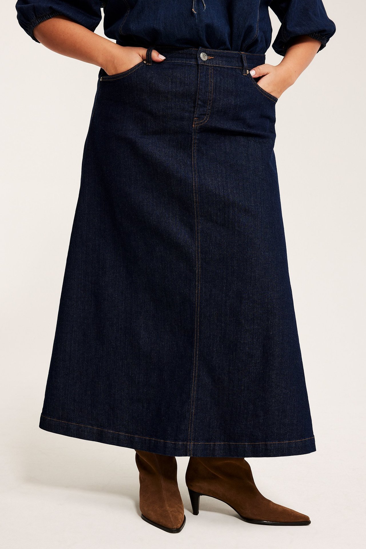 Frontvy av en kvinna i en mörkblå A-linje jeanskjol, en mörk topp och bruna mockakängor.