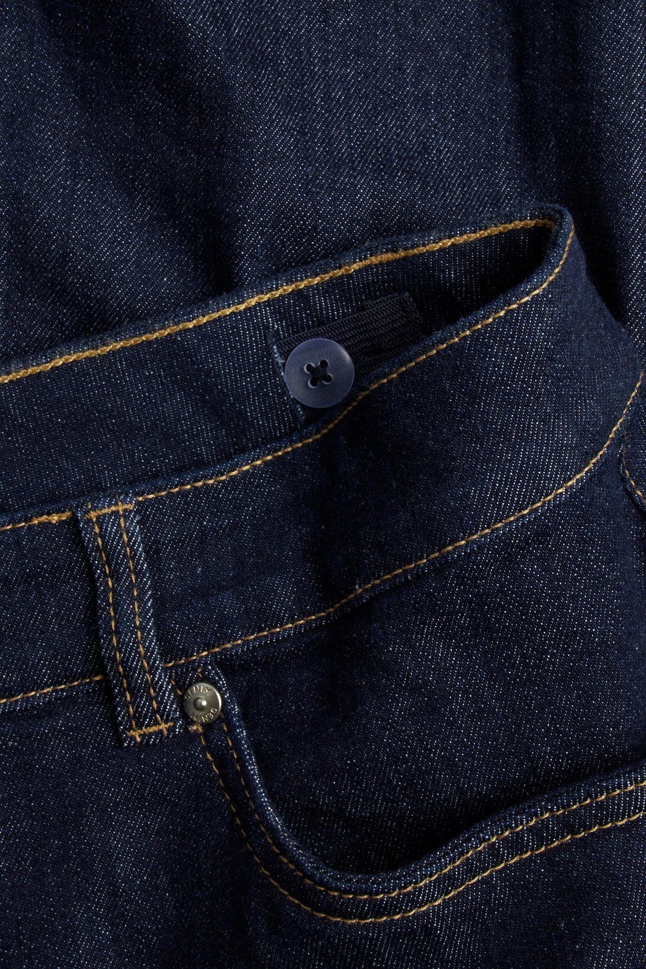 Zbliżenie: ciemnoniebieska jeansowa spódnica A-linii, z guzikiem, regulowaną gumką i żółtymi szwami.