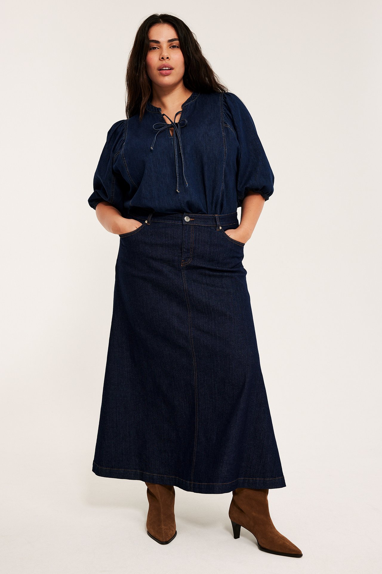Kvinna i lång mörkblå jeanskjol i A-linje, mörkblå blus med puffärmar och bruna mockakängor. Framifrån.