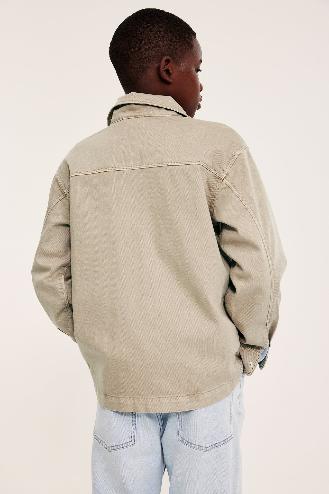Bakifrån, ett barn i en lös beige twill-overshirt och ljusblå jeans.