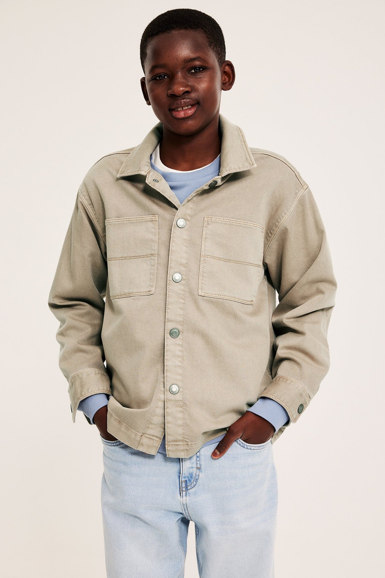 Edestäpäin: Lapsi beigessä puuvillatwill-päällistakissa painonapeilla, vaaleansinisen paidan ja vaaleansinisten farkkujen päällä.