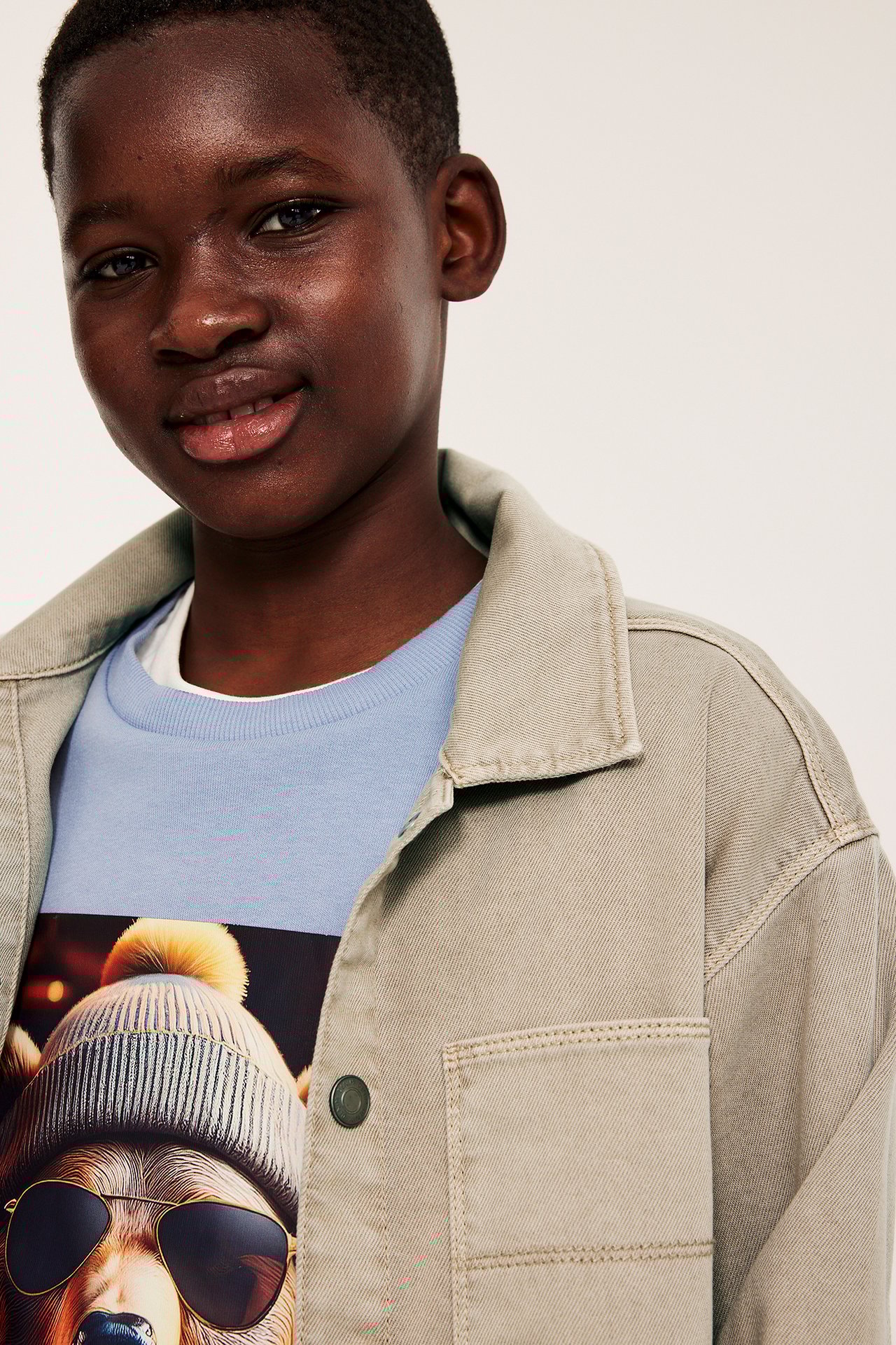 Närbild: Kid i öppen beige twill-overshirt med tryckknappar över en ljusblå grafisk t-shirt.