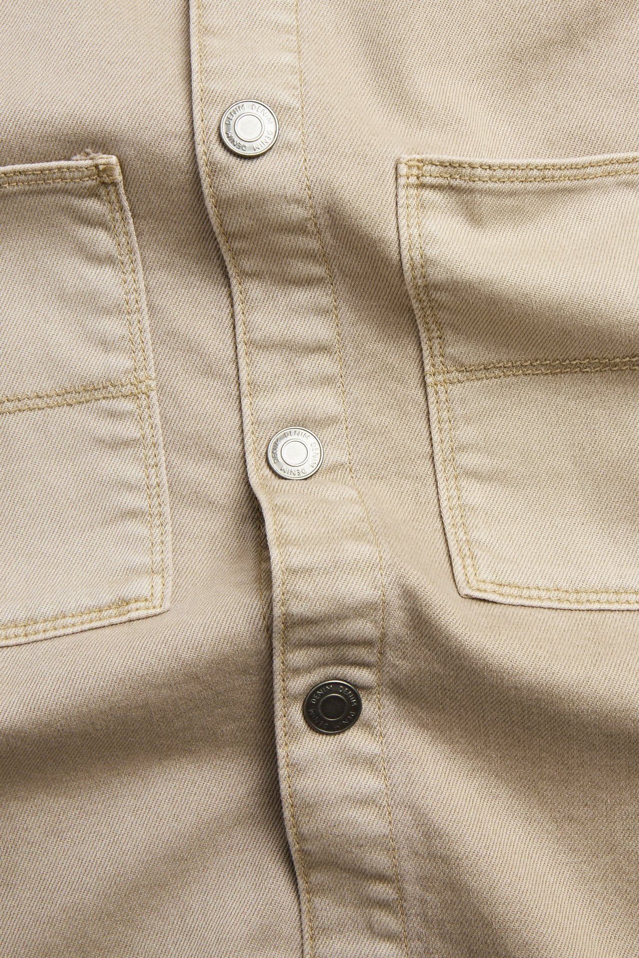 Närbild, framifrån, av en beige twill-overshirt i bomull med jeansknappar och bröstfickor.
