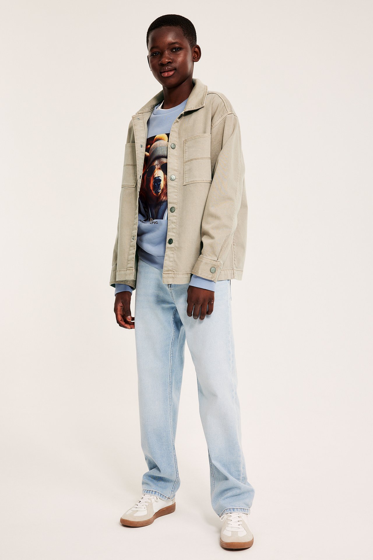 Edestäpäin: Lapsi beigessä twill-ylipaidassa, vaaleansinisessä collegepaidassa, vaaleansinisissä farkuissa ja tennareissa.