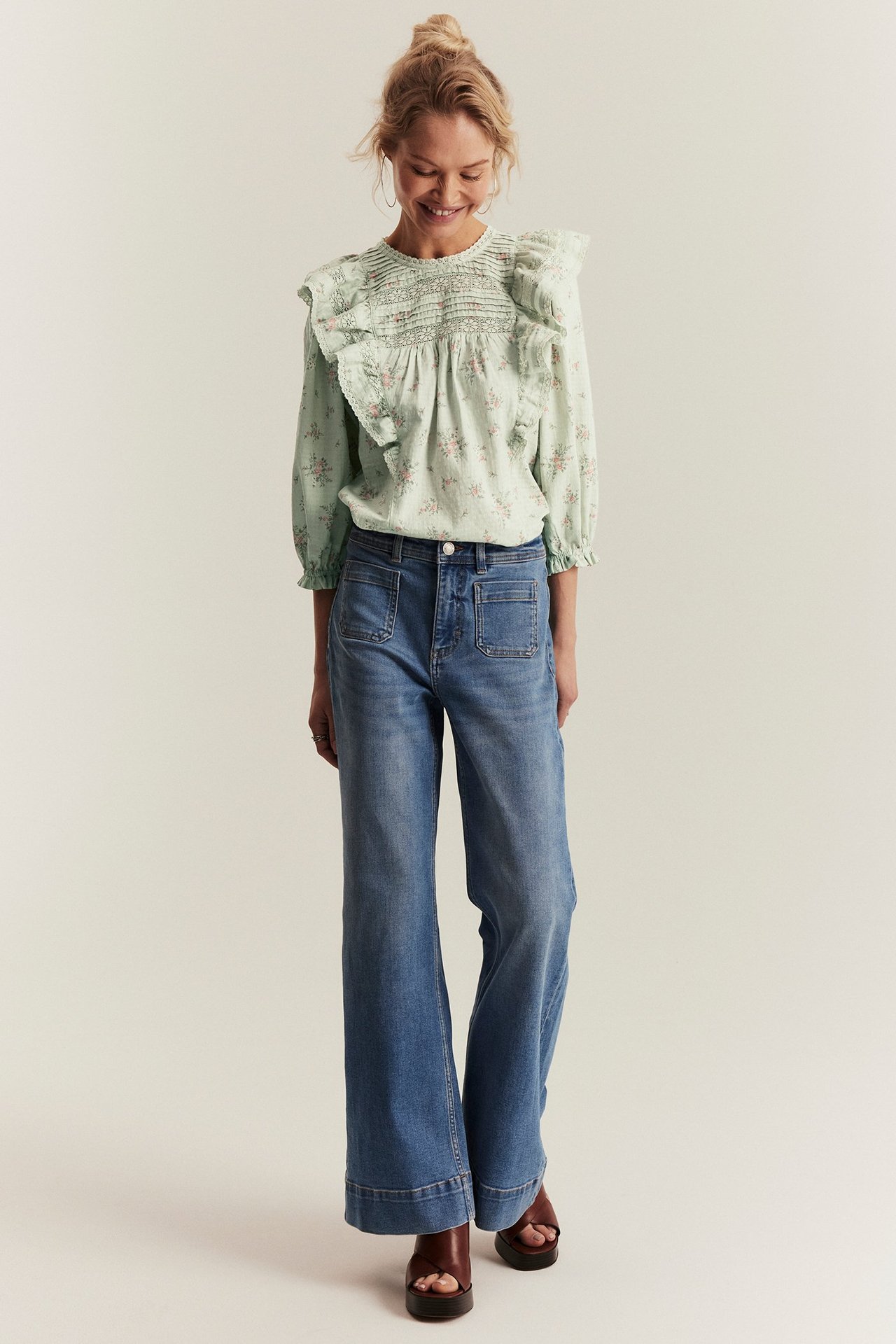 Edestäpäin: nainen vaaleanvihreässä kukkaisessa pitsipuserossa, smokkirypytyksellä, röyhelöillä, sinisissä leveissä farkuissa ja ruskeissa sandaaleissa.
