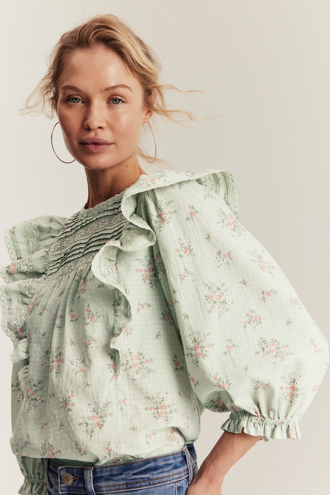 Edestäpäin nainen vaaleanvihreässä kukkakuosisessa puuvillapuserossa, jossa röyhelöitä, pussuhihat, farkut ja vannekorvakorut.