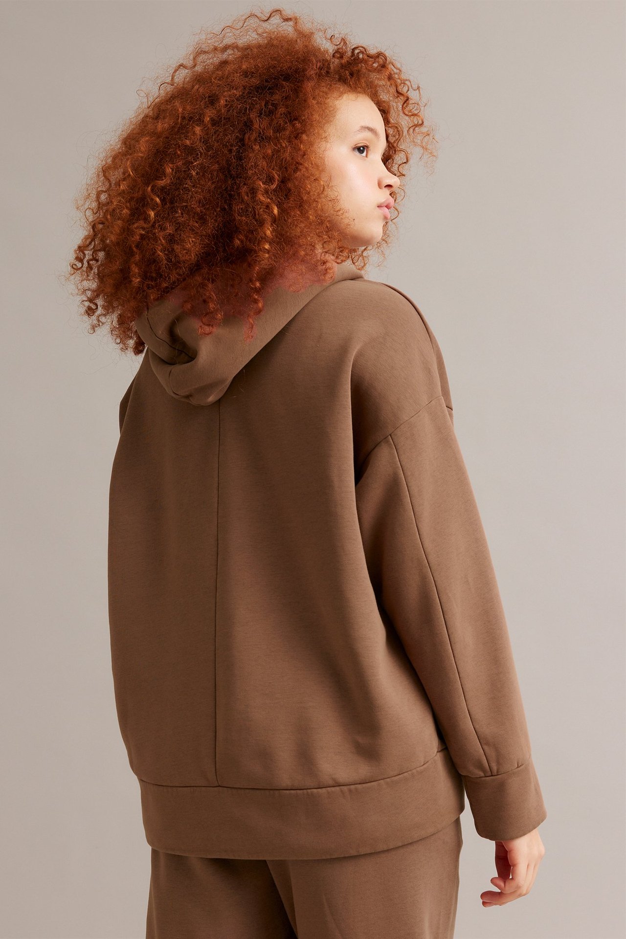 Takakuva naisesta, jolla on väljä, ruskea puuvillasekoitehuppari hupulla ja collegehousut.