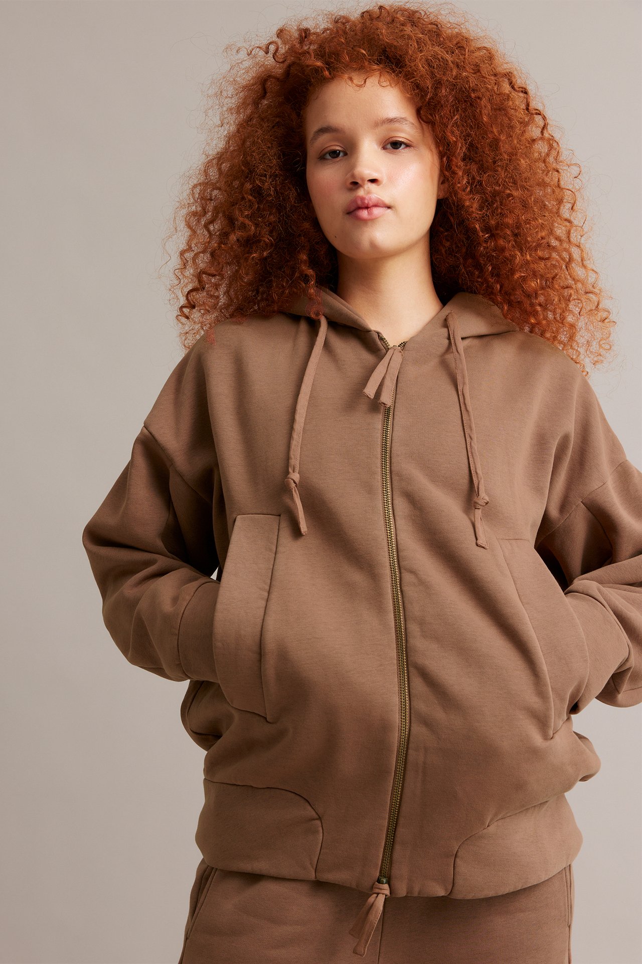 Edestä, nainen löysässä ruskeassa vetoketjullisessa hupparissa, kädet taskuissa, yllään yhteensopivat housut.