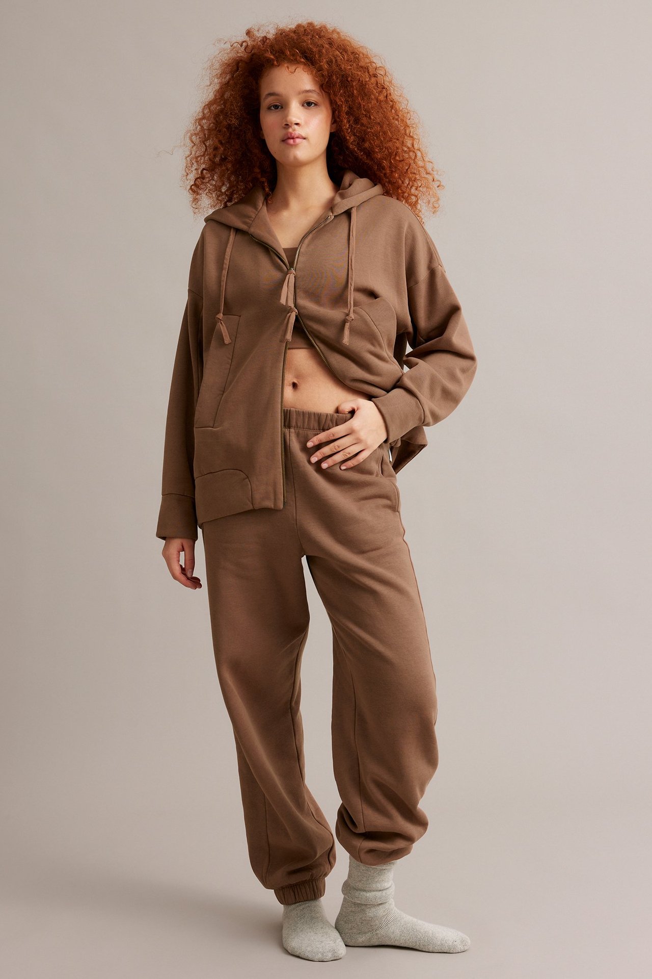 Edestä: nainen ruskeassa vetoketjuhupparissa, yhteensopivissa collegehousuissa ja vaaleanharmaissa sukissa.