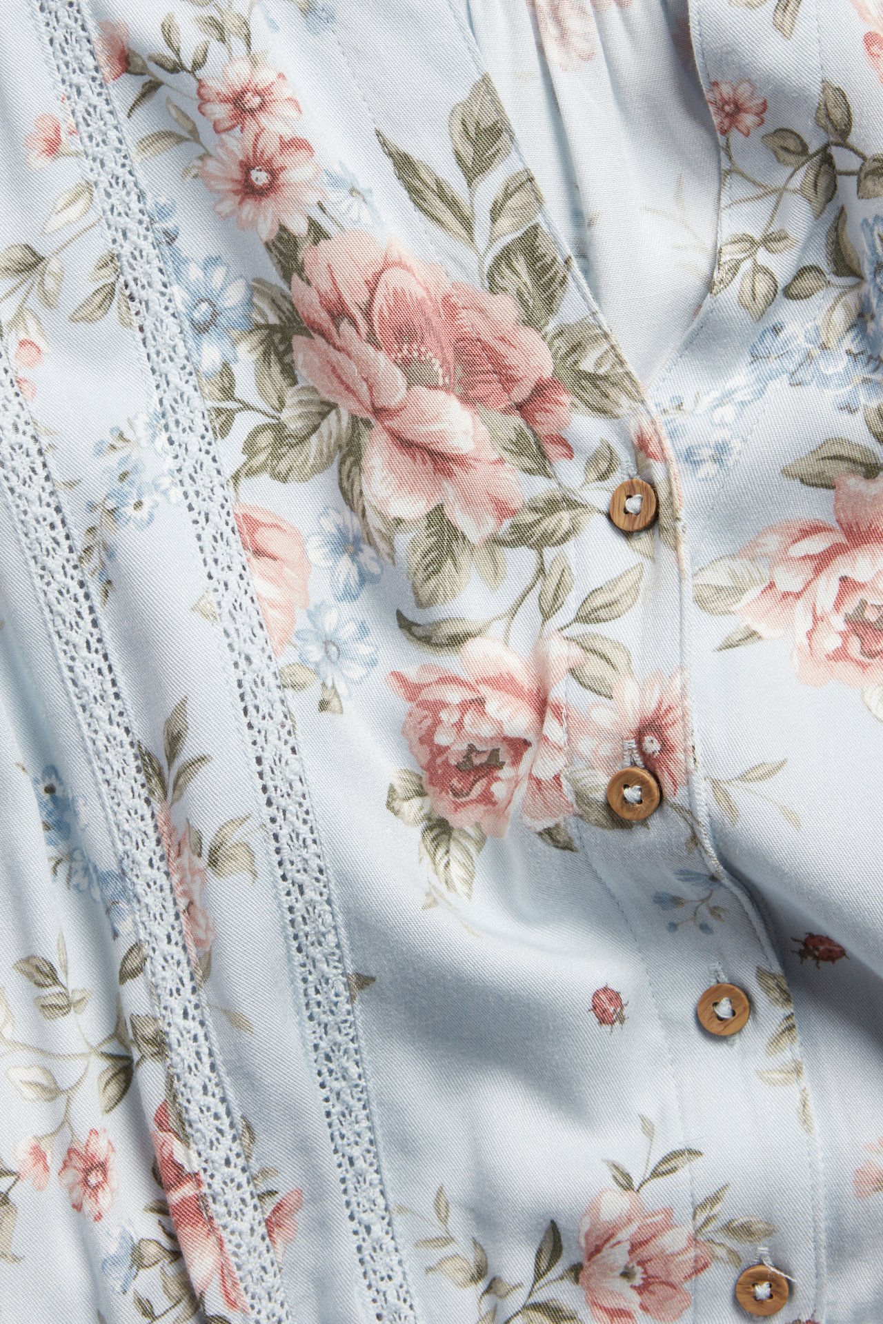 Nærbilde av en lyseblå blomstret viskosekjole med blondeinnfellinger og små treknapper.