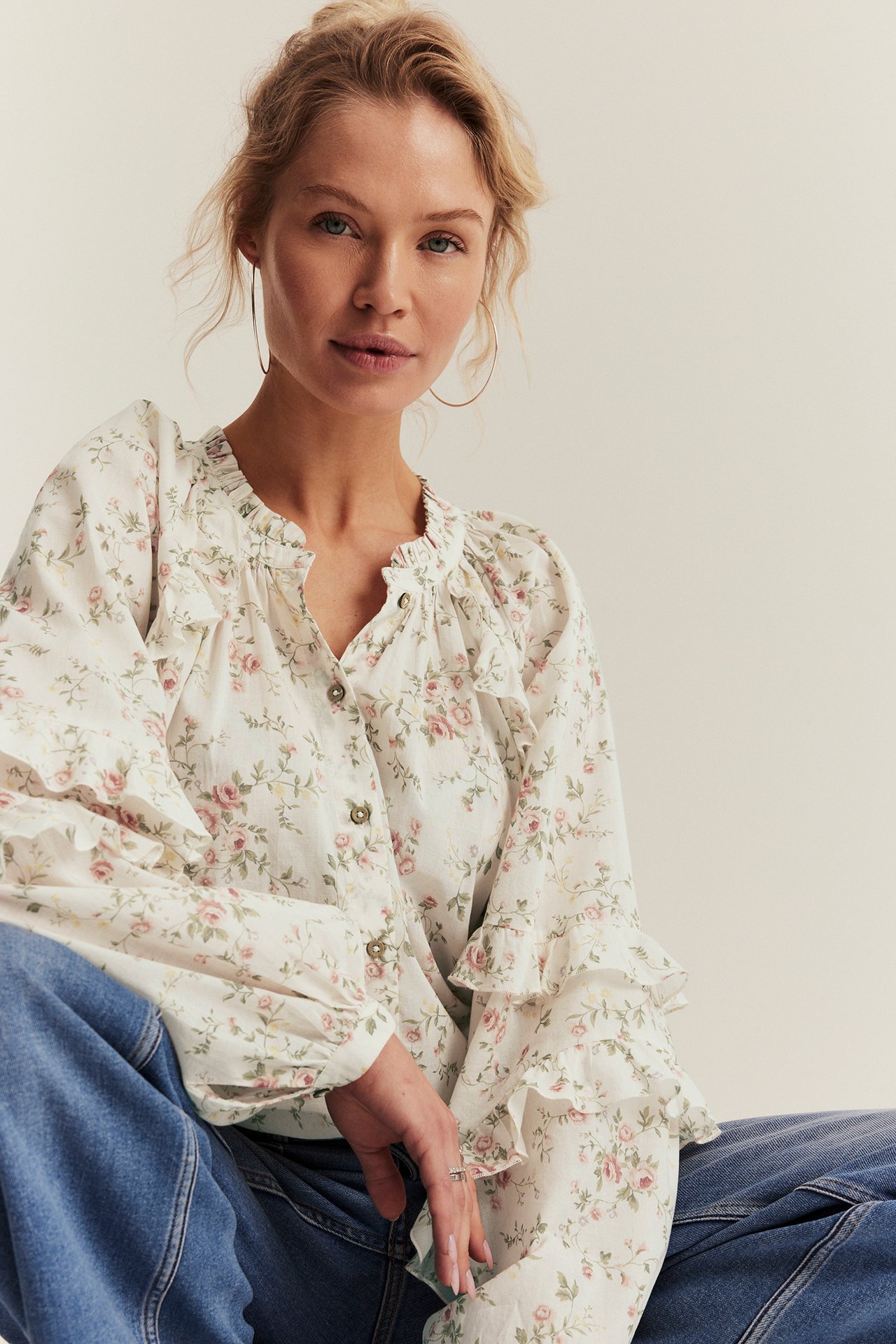 Frontvisning av kvinne i offwhite blomstret bomullsbluse med volanger, knapper og blå jeans.