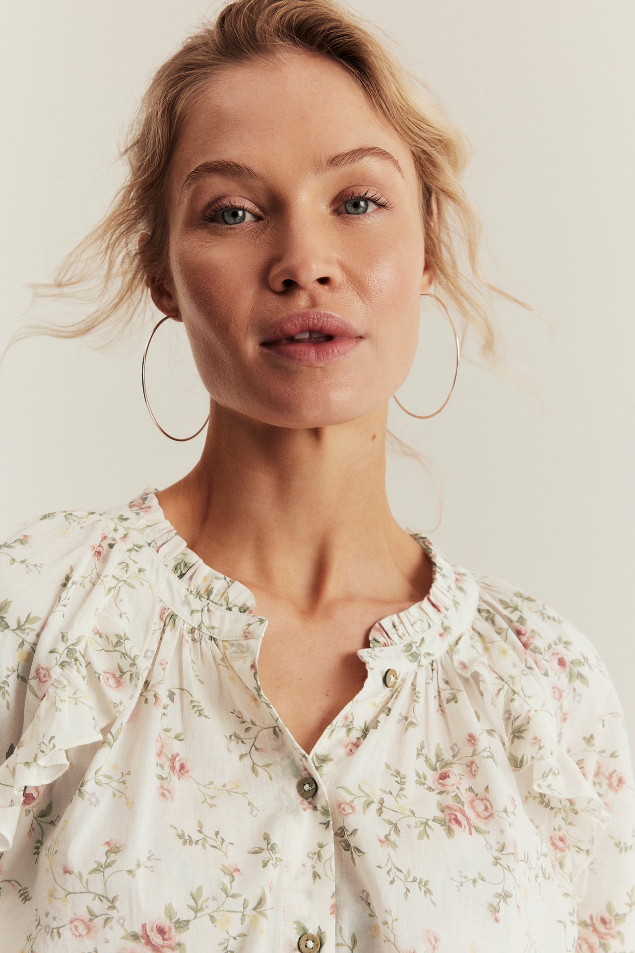 Kvinne har på seg off-white blomstret bomullsbluse med rysjer, knapper foran og store ringøreringer.