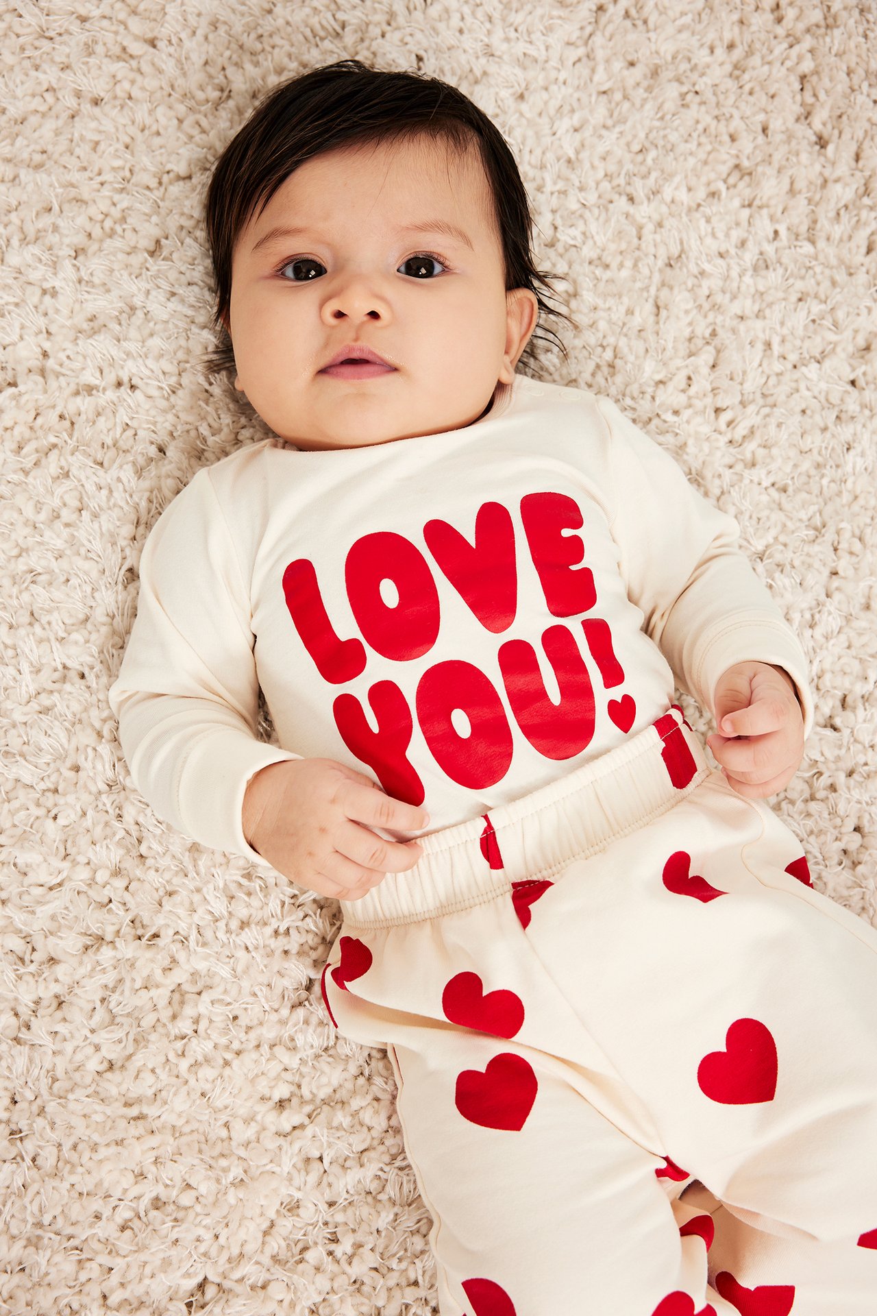 Forfra: En baby i offwhite langermet body med rødt 'LOVE YOU!'-trykk og matchende hjerteprintede bukser.