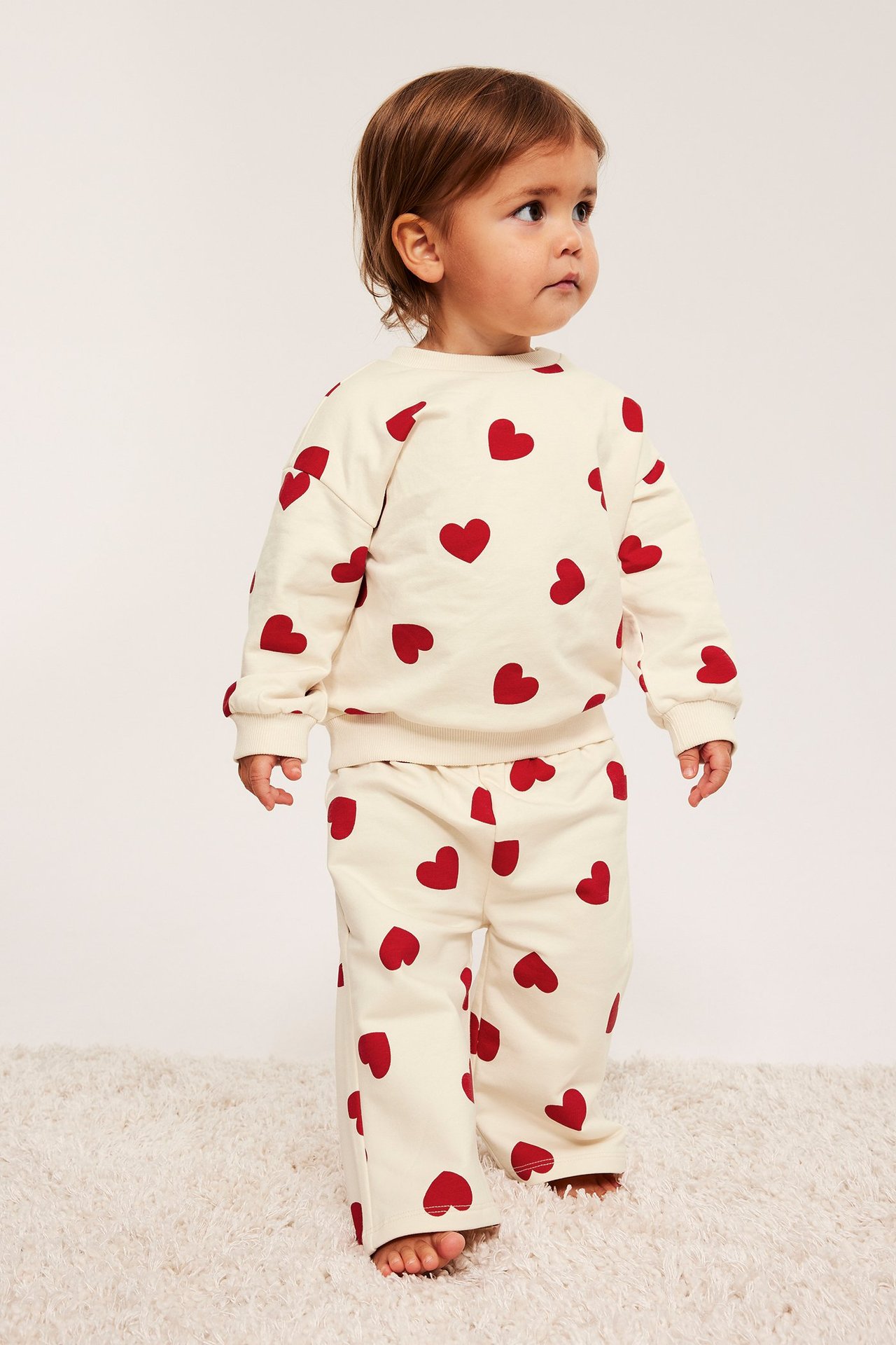 Edestä, vauva luonnonvalkoisessa college-setissä, punaisilla sydämillä, paita ja suoralahkeiset housut.
