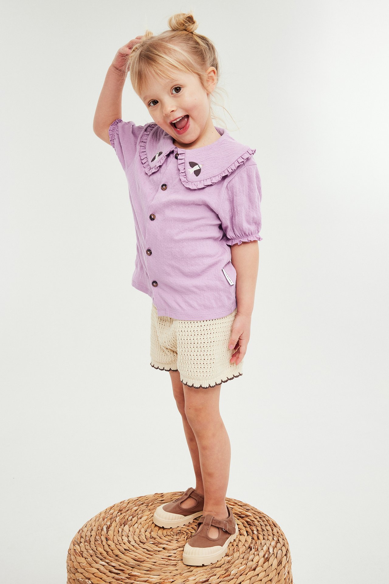 Barn i lila krinklad blus med broderad krage, beige virkade shorts och bruna skor.
