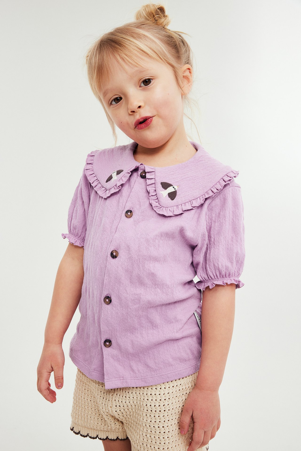 Barn bär en ljuslila krinklad blus med volangkrage, fågelbrodyr, knappar och beiga virkade shorts.