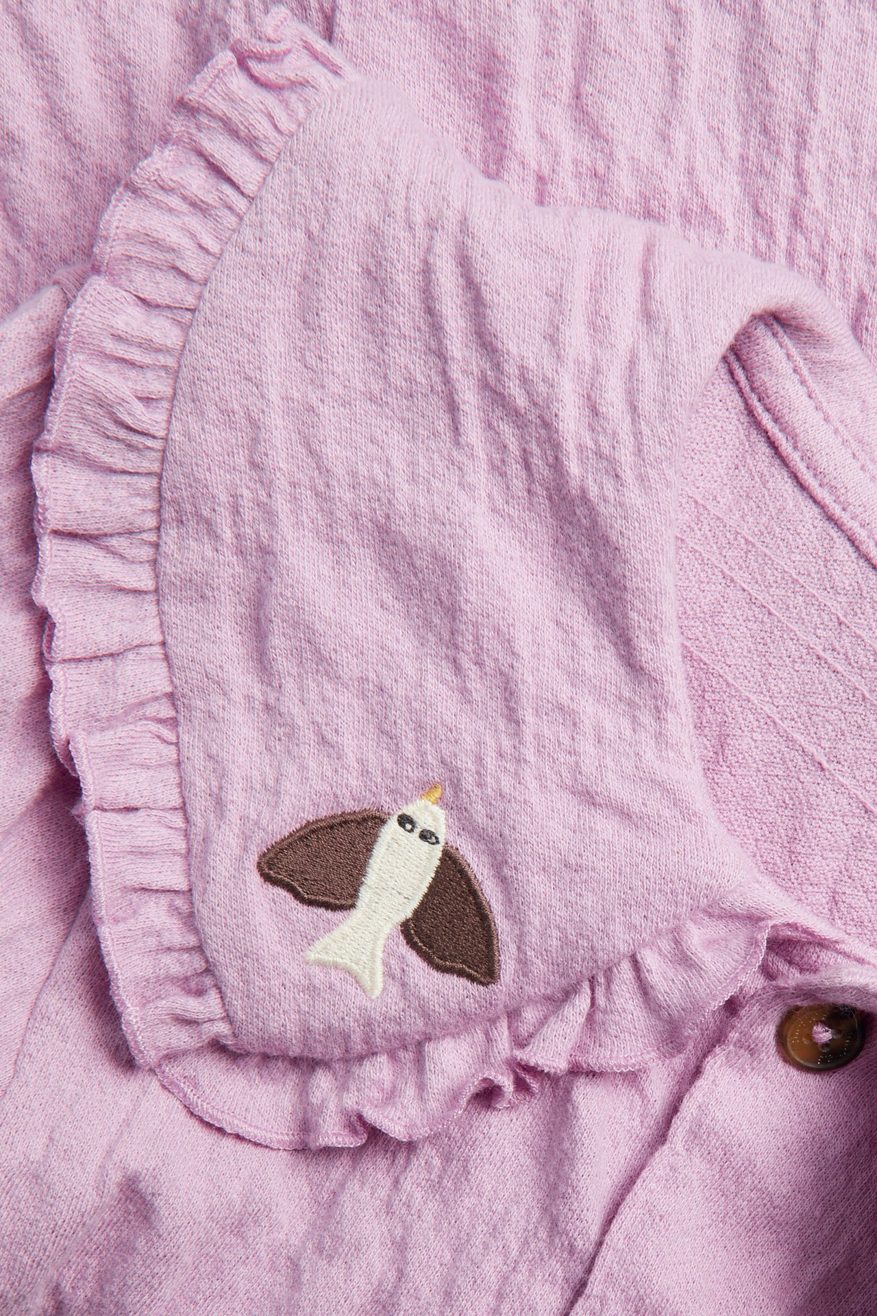 Zbliżenie różowej, kreszowanej bluzki dziecięcej z dzianiny, z falbankowym kołnierzem, haftem ptaka i brązowymi guzikami.