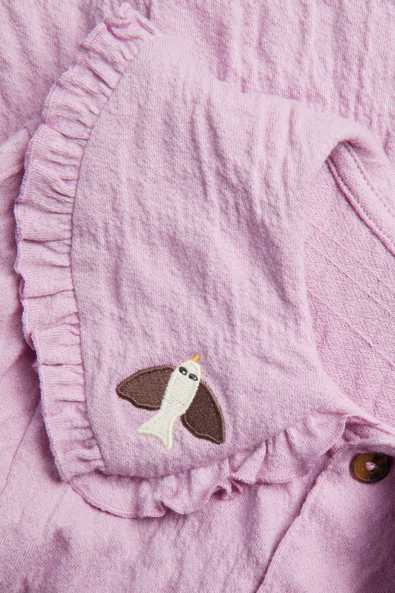 Zbliżenie różowej, kreszowanej bluzki dziecięcej z dzianiny, z falbankowym kołnierzem, haftem ptaka i brązowymi guzikami.