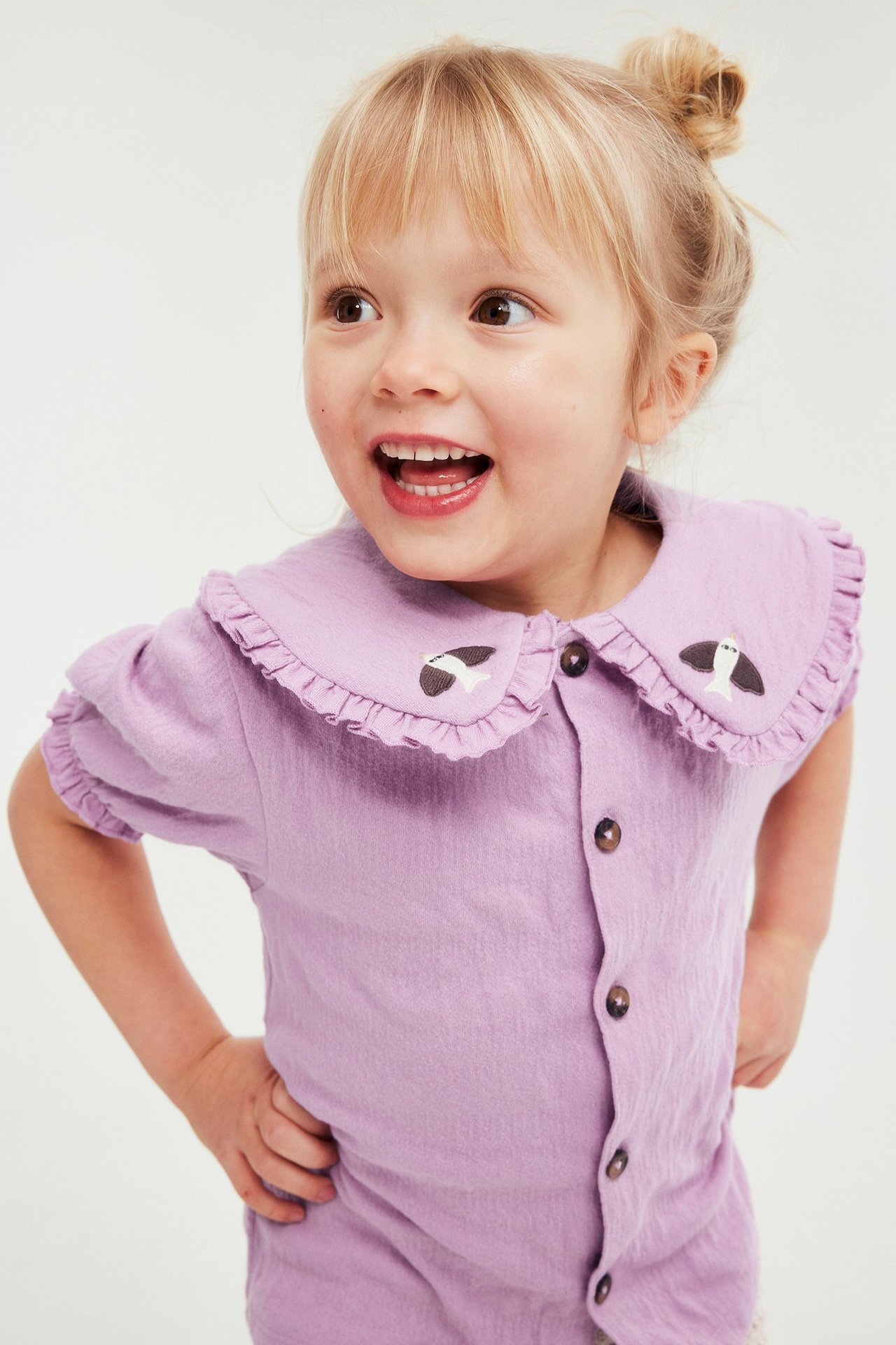 Lapsi liilassa, rypytetyssä puserossa, jossa röyhelökaulus, kirjonta ja ruskeat napit, edestäpäin.