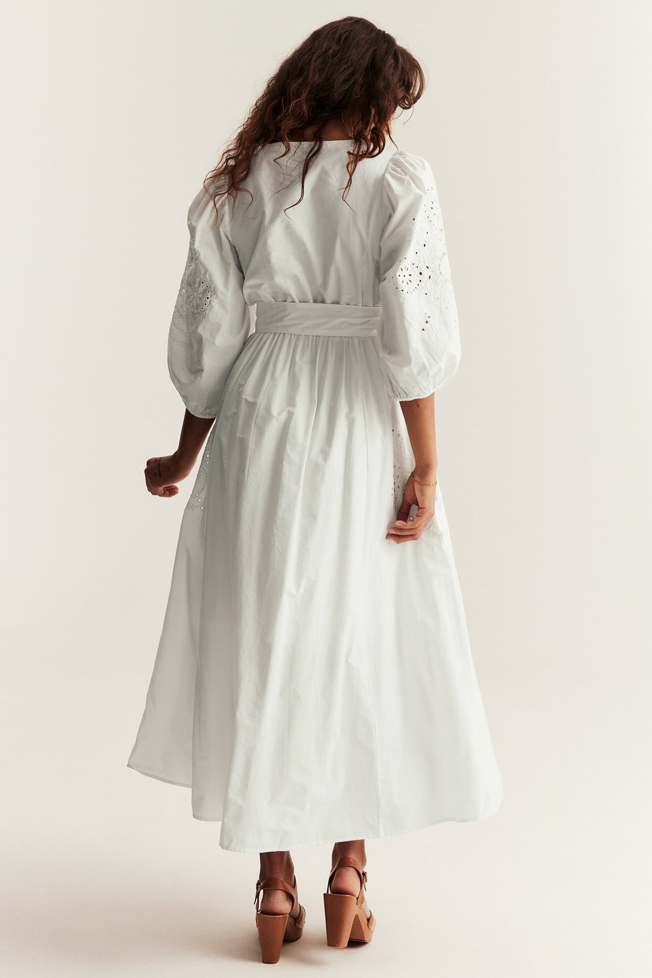 Baksidan av en kvinna i offwhite broderad bomullsklänning med puffärmar och bruna kilklackssandaler.