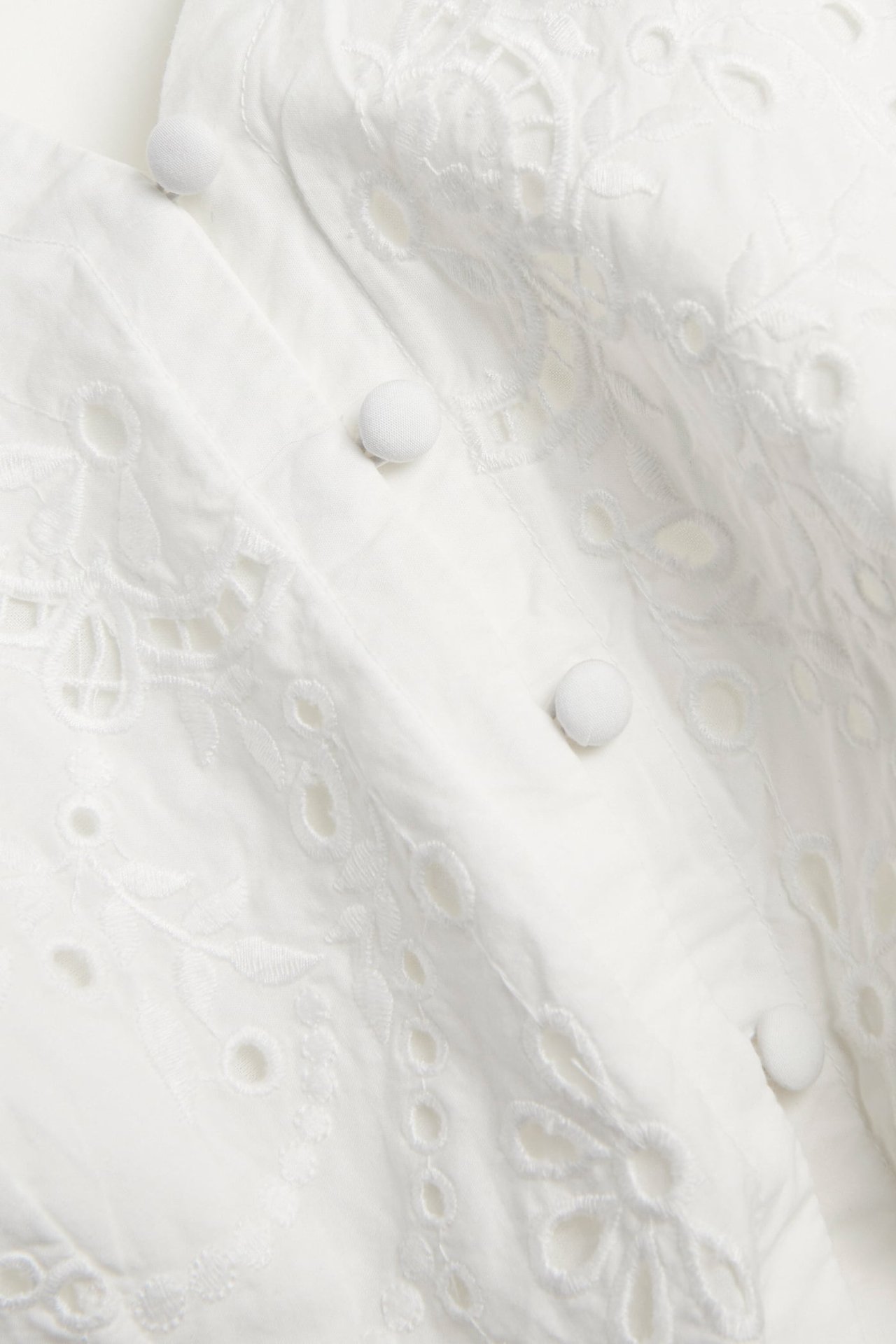 Närbild av offwhite bomullsklänning med broderier, hålmönster och tygklädda knappar framtill.