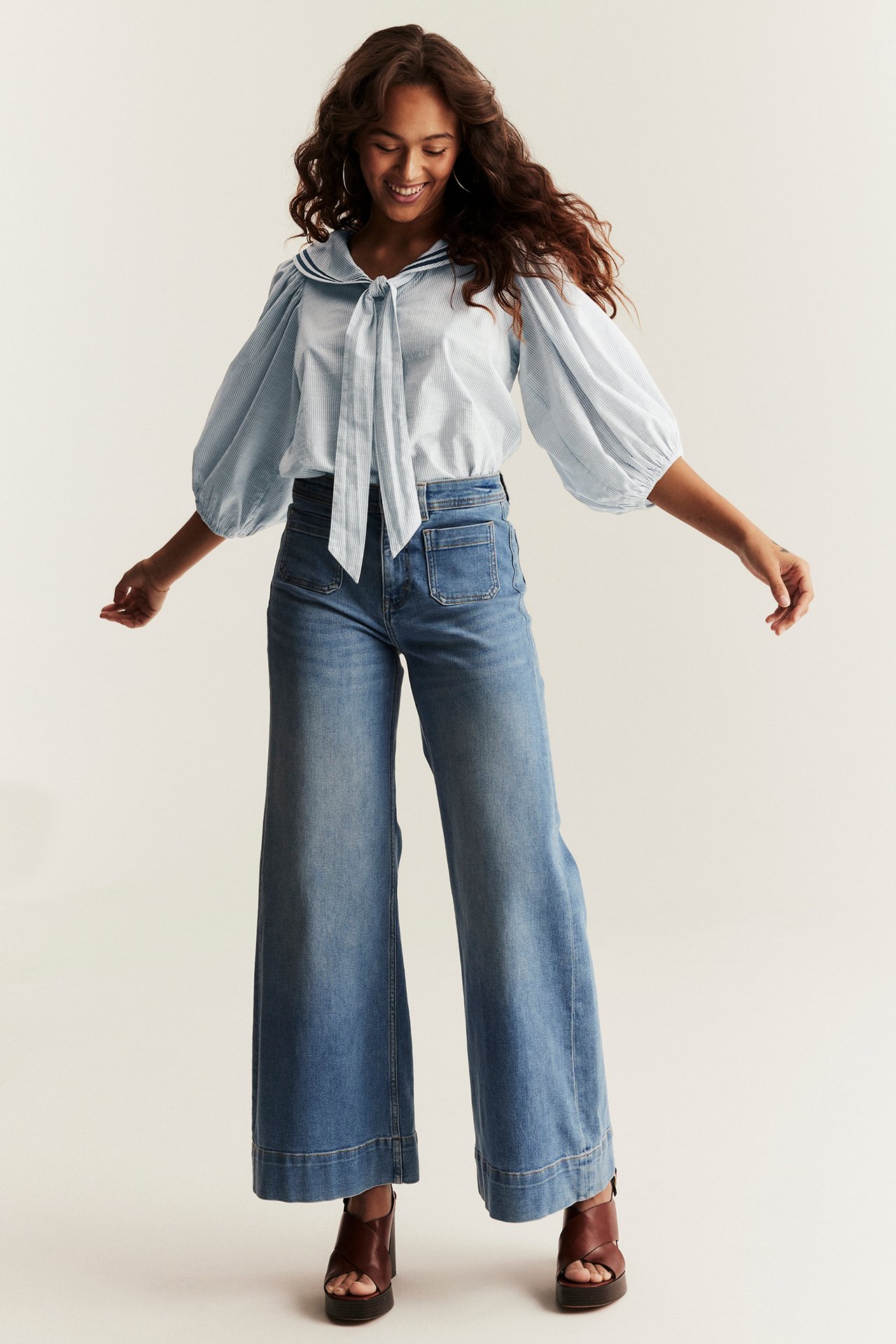Kvinna bär ljusblå randig seglarblus med knytband, puffärmar, blå vida jeans, bruna platåsandaler.