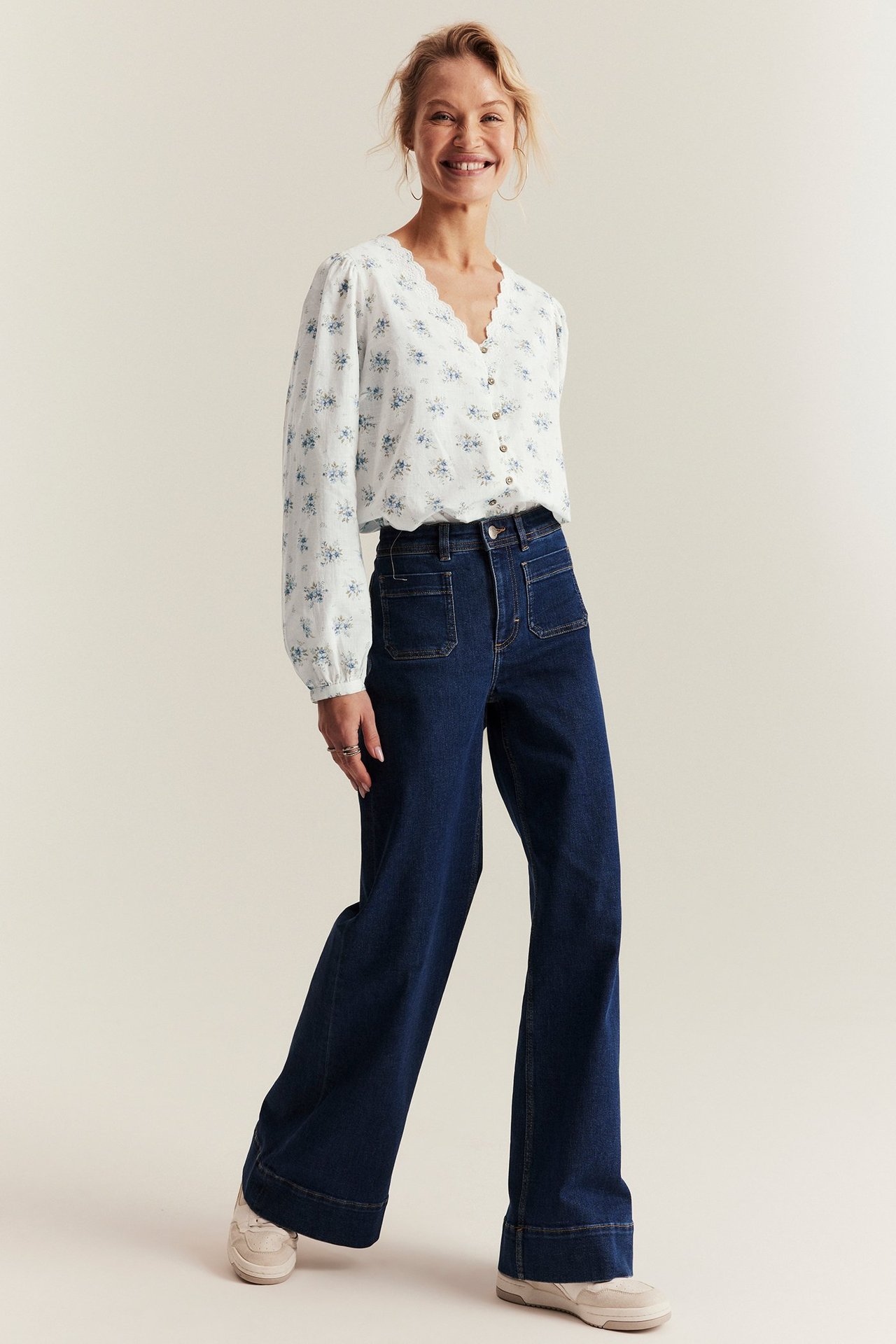 Nainen luonnonvalkoisessa kukallisessa V-aukko puserossa, pussihihat, tummansiniset farkut, vaaleat lenkkarit.