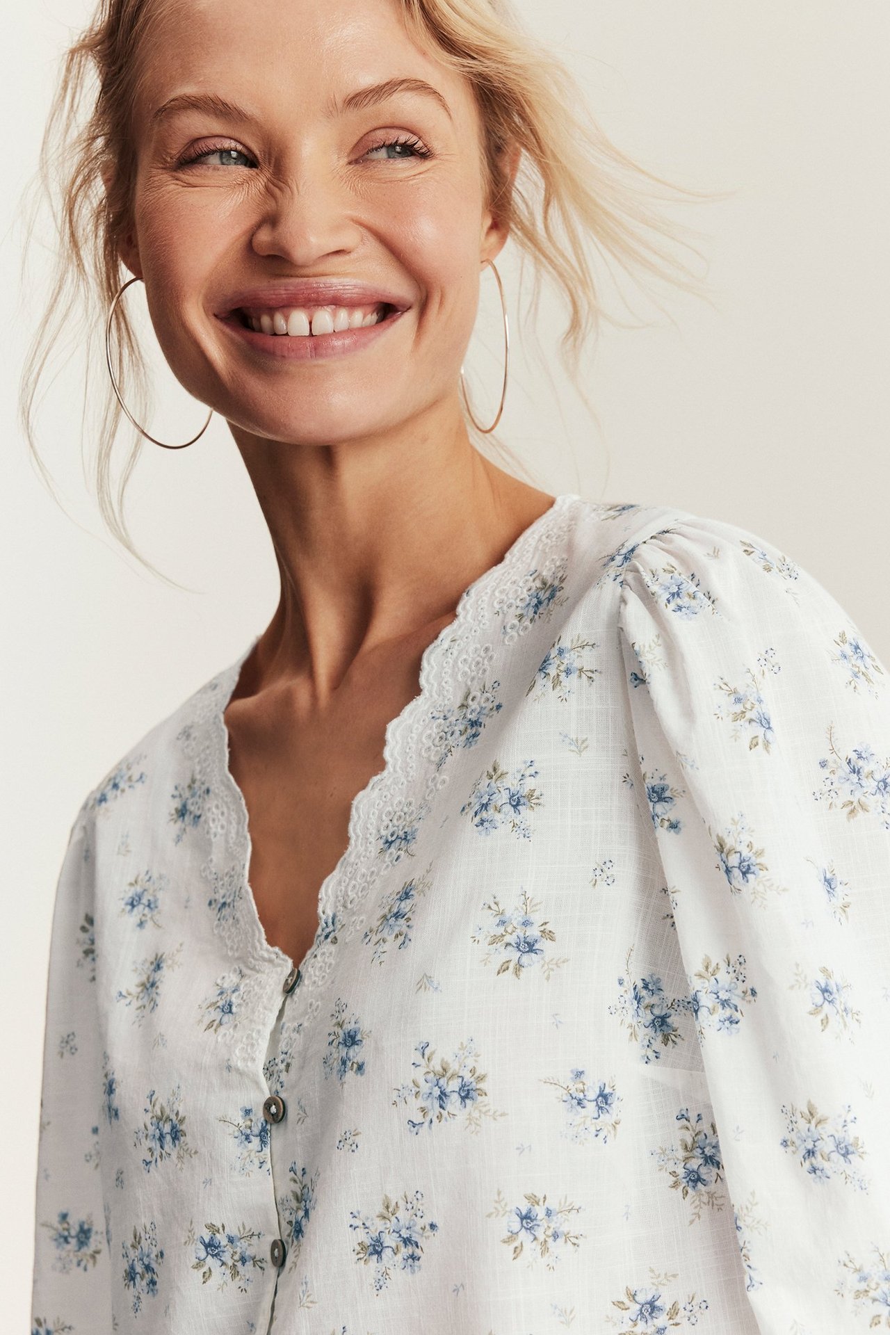 Lähikuva: Nainen pukeutuneena luonnonvalkoiseen kukkakuvioiseen puuvillapuseroon, jossa on kirjailtu V-pääntie ja puffihihat, kultaiset vannekorvakorut.