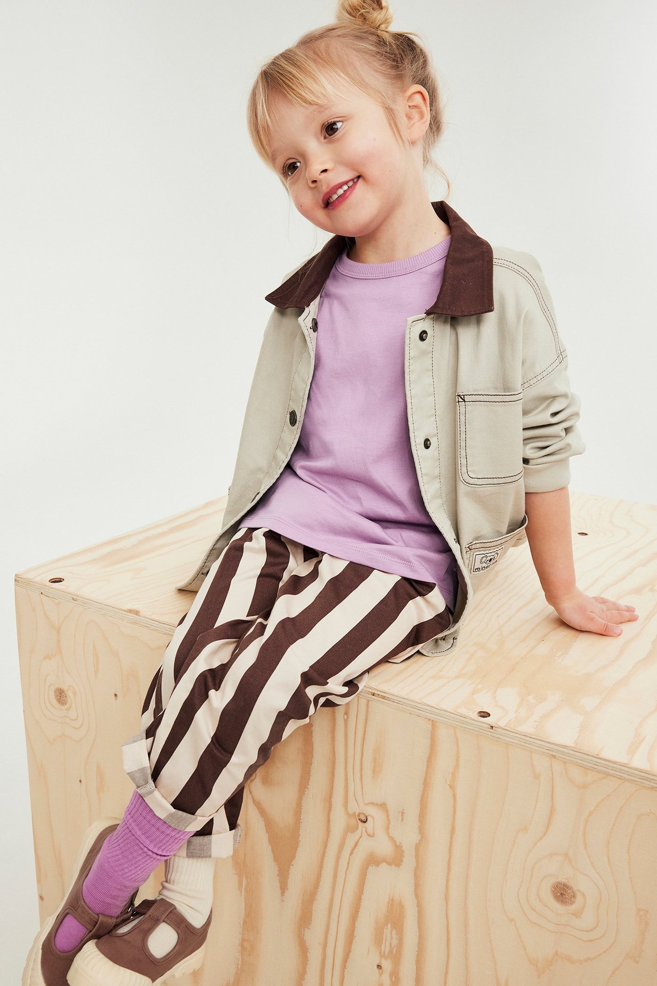 Forside: Et barn i en lyslilla t-skjorte, beige jakke, stripete bukser, lilla sokker og brune sko.