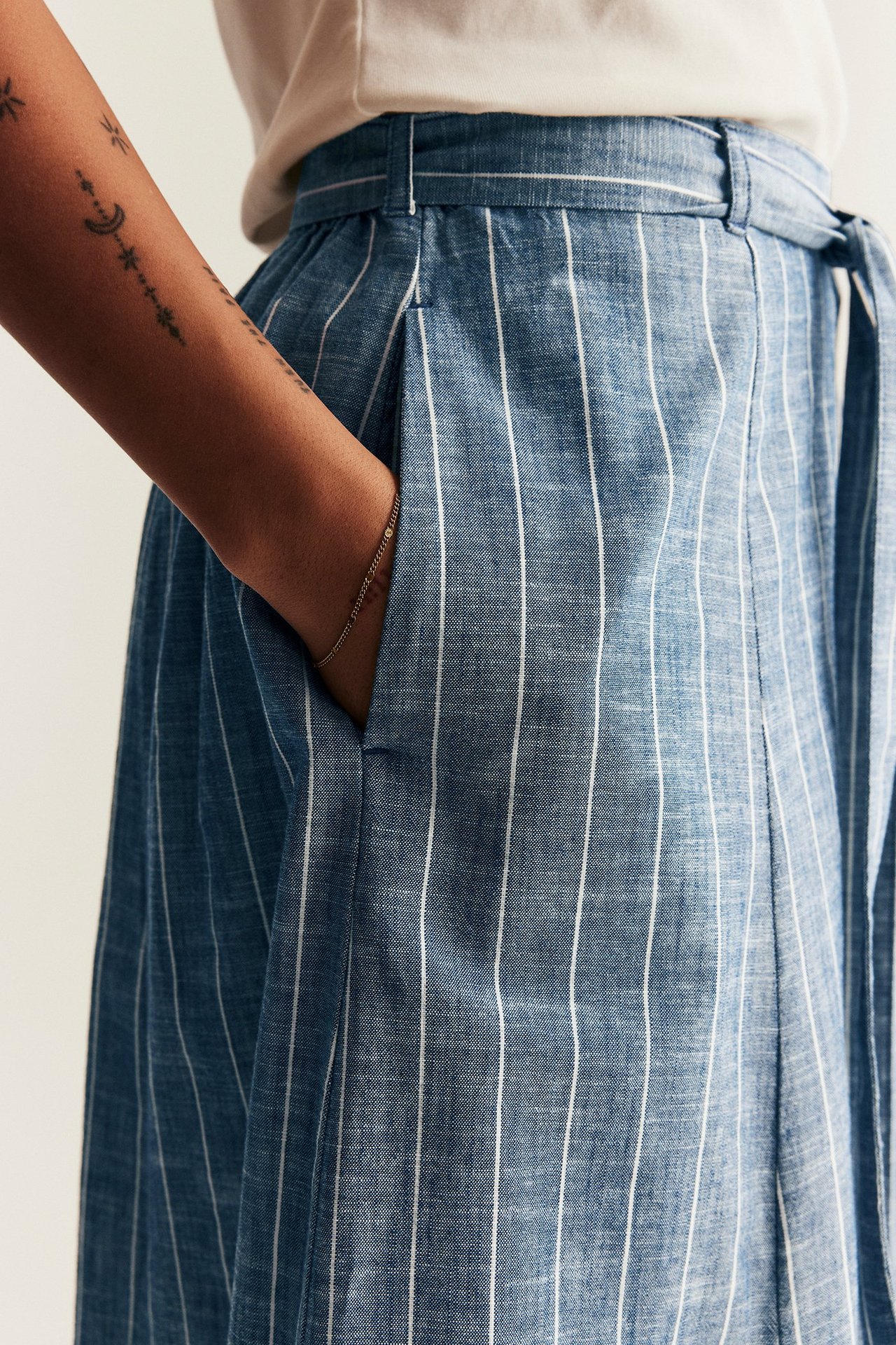 Lähikuva naisesta sinivalkoraidallisessa kudotussa hameessa, jossa sivutasku, solmittava vyö, ja vaalea t-paita sekä kultarannekoru.