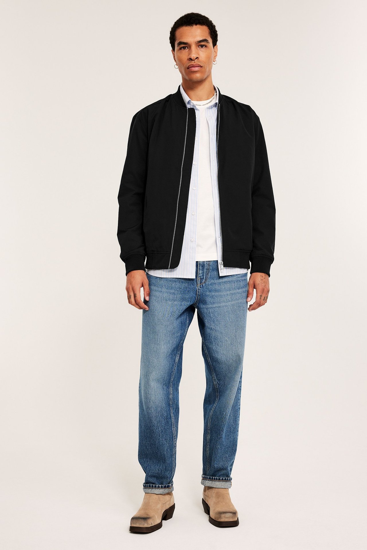 Forfra: mann i svart bomberjakke over stripet skjorte, blå jeans og beige boots.