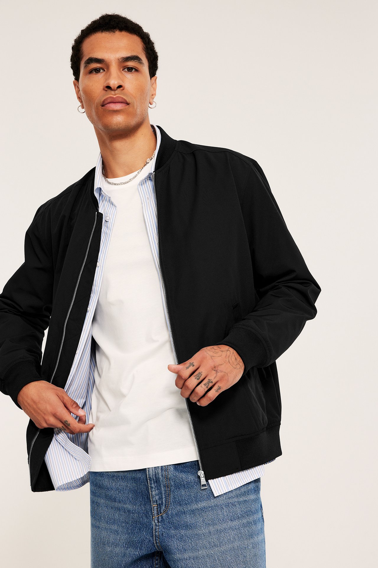 Frontutsikt: mann i åpen svart bomberjakke, hvit t-skjorte, lyseblå stripet skjorte og blå jeans.