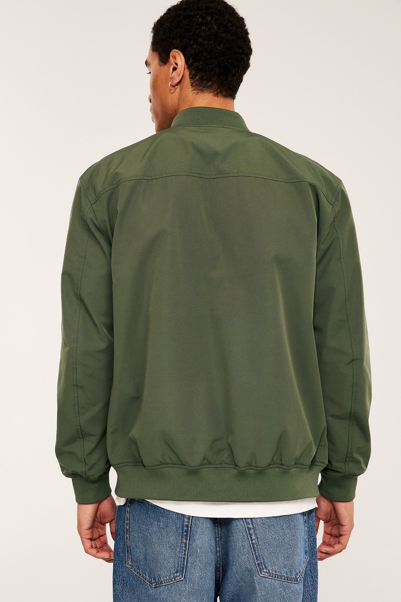 Mann, bakfra, i en mørkegrønn bomberjakke med ribbestrikkede detaljer, over en hvit T-skjorte og blå jeans.
