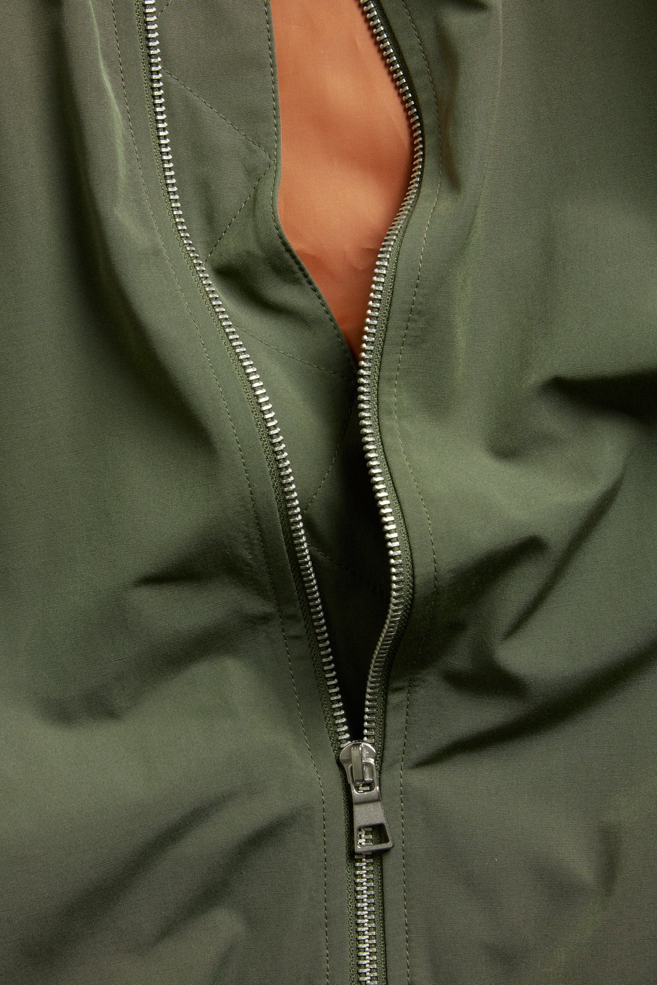 Nærbilde forfra av en mørkegrønn bomberjakke med delvis åpen sølvfarget metallglidelås og oransje fôr.