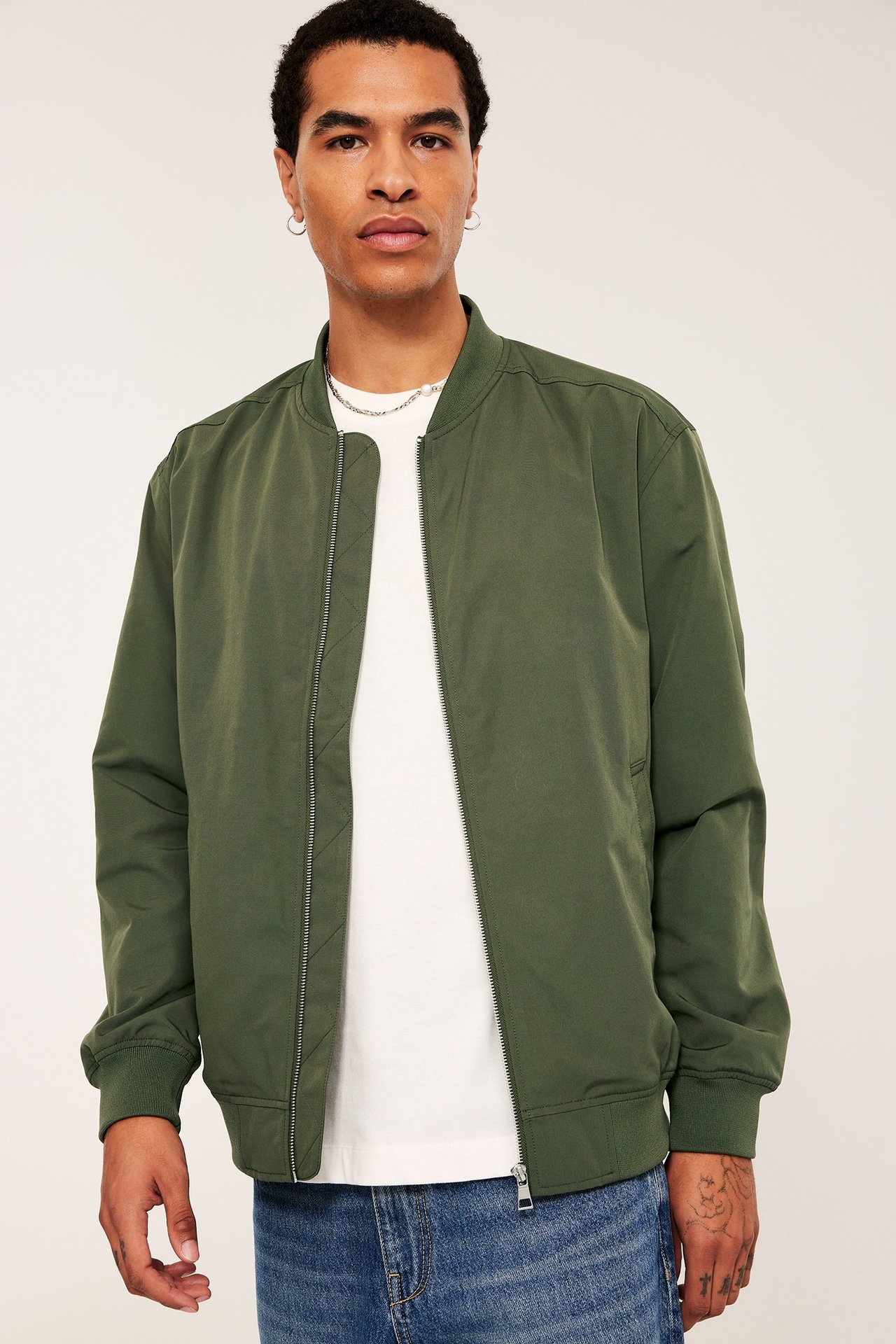 Edestä: Mies avonaisessa tummanvihreässä bomber-takissa, valkoisessa t-paidassa ja sinisissä farkuissa.