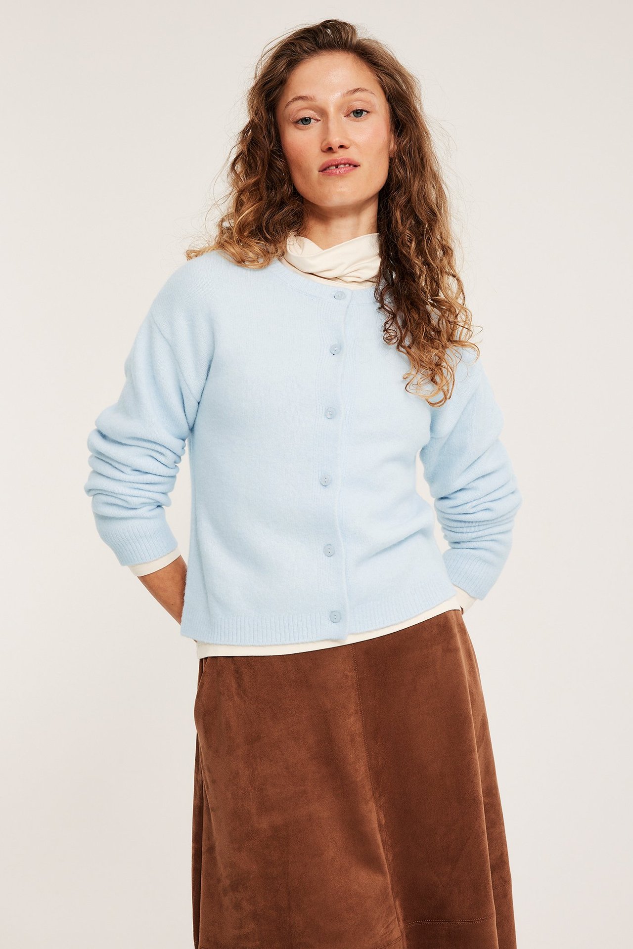 Edestä: Nainen vaaleansinisessä hienoneuletakissa nappikiinnityksellä, kermanvärisen poolopaidan ja ruskean hameen päällä.