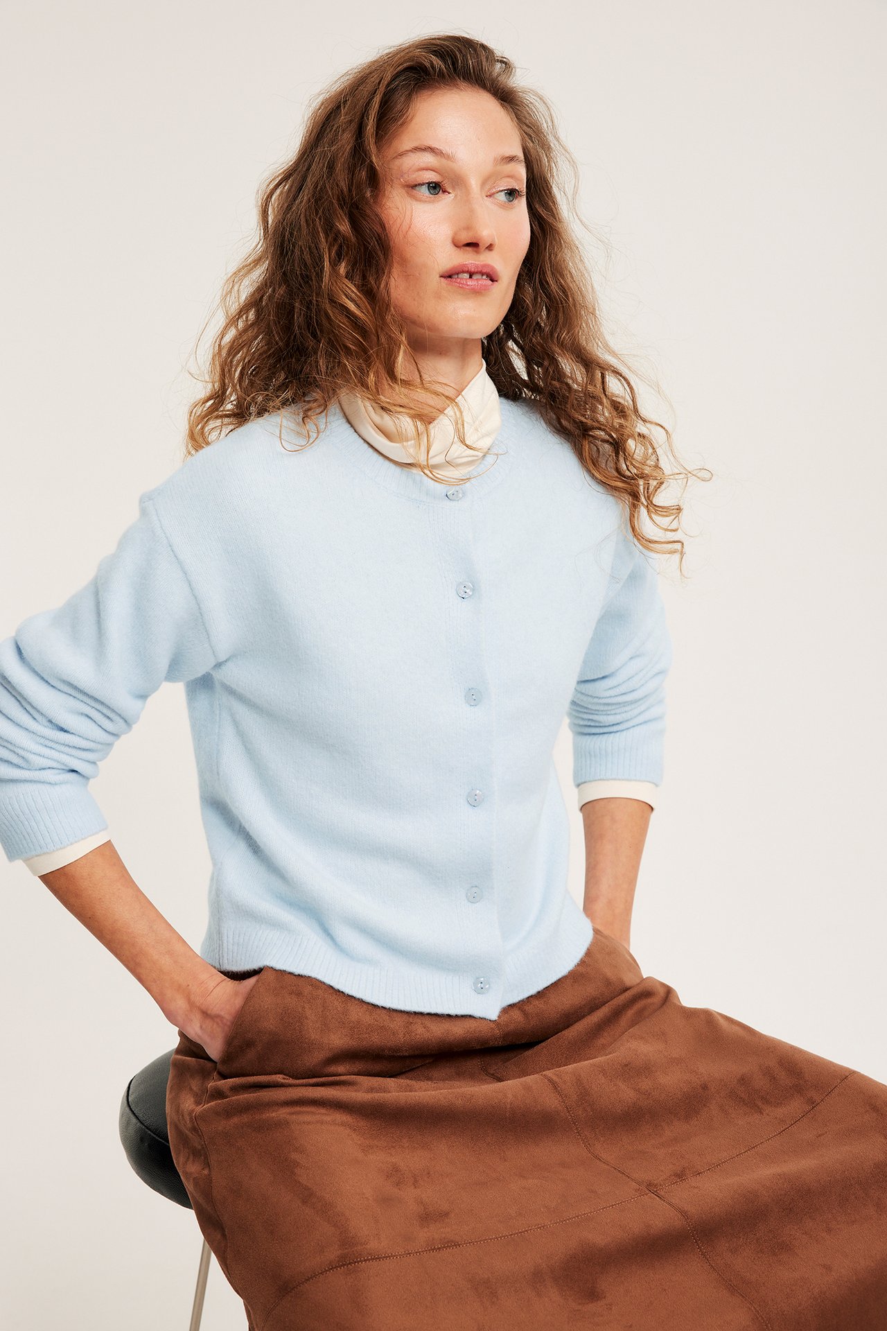 Naisen edestä kuvattu vaaleansininen ohut neuletakki nappikiinnityksellä valkoisen paidan ja ruskean hameen päällä.