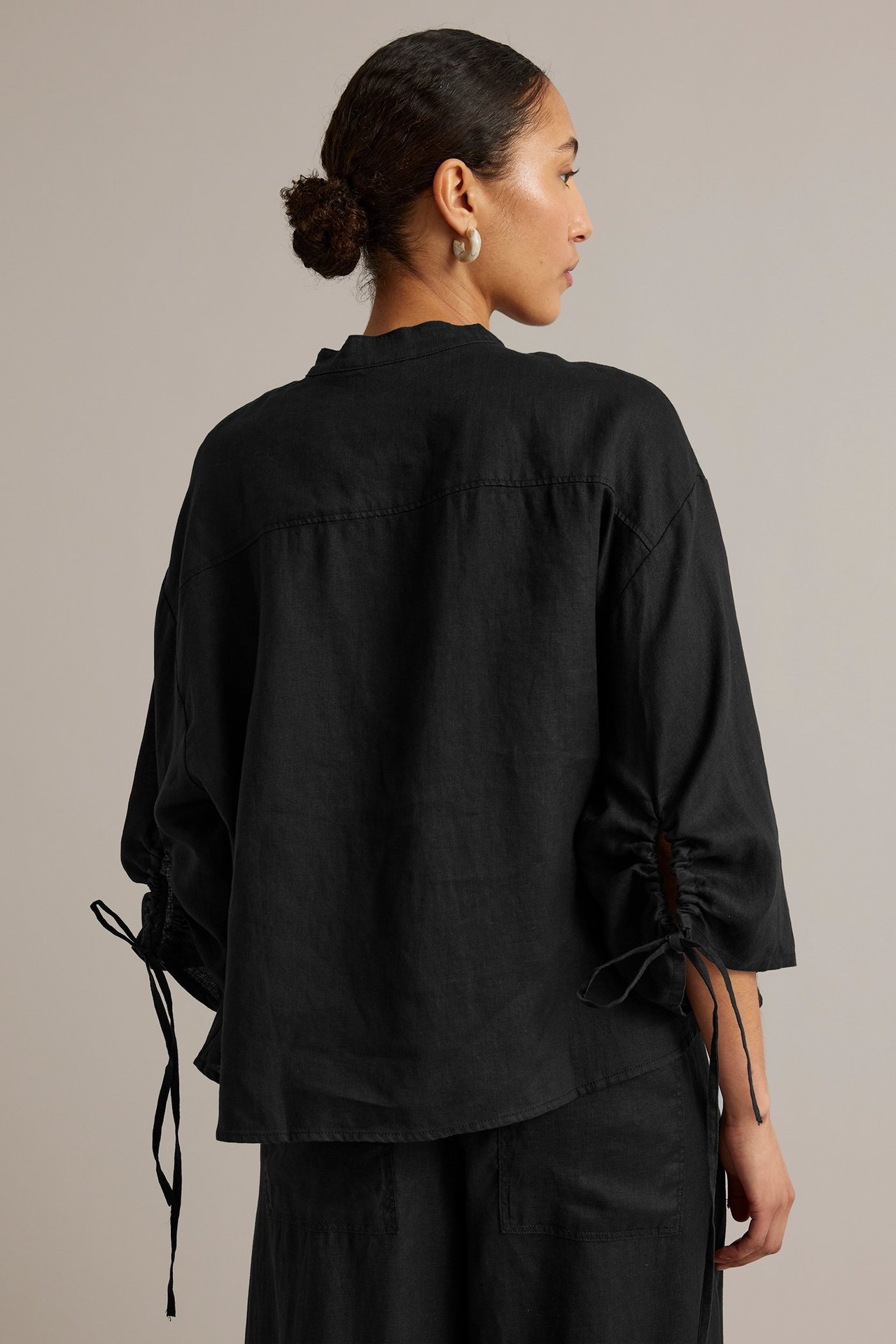 Ryggbilde av en kvinne i svart linbluse med rynkede ermer, mansjettbånd og svarte bukser.