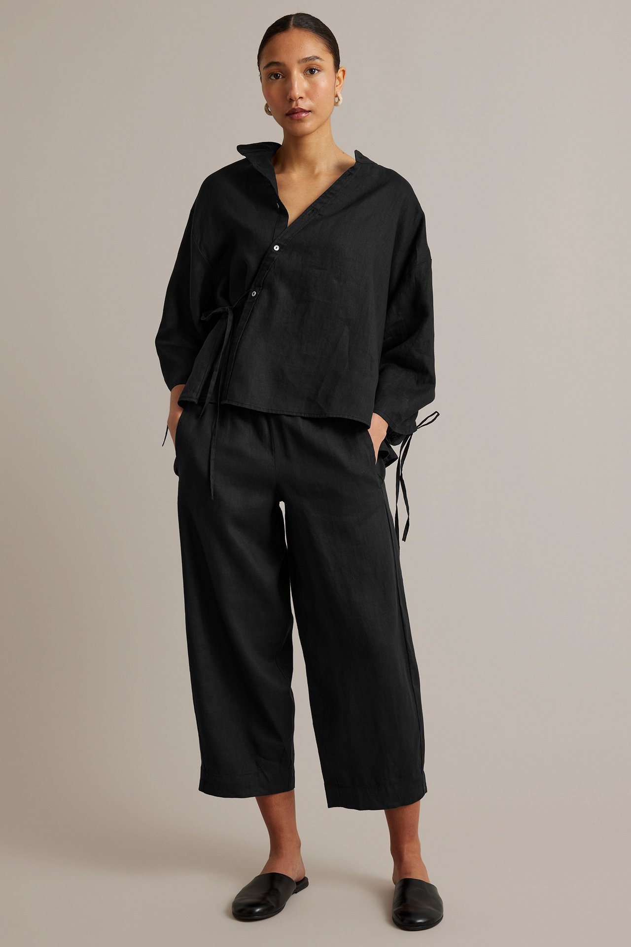 Edestäpäin: Nainen yllään musta pellavapusero vinonapilla, vyötärönauhalla, leveälahkeiset housut ja mustat muulit.