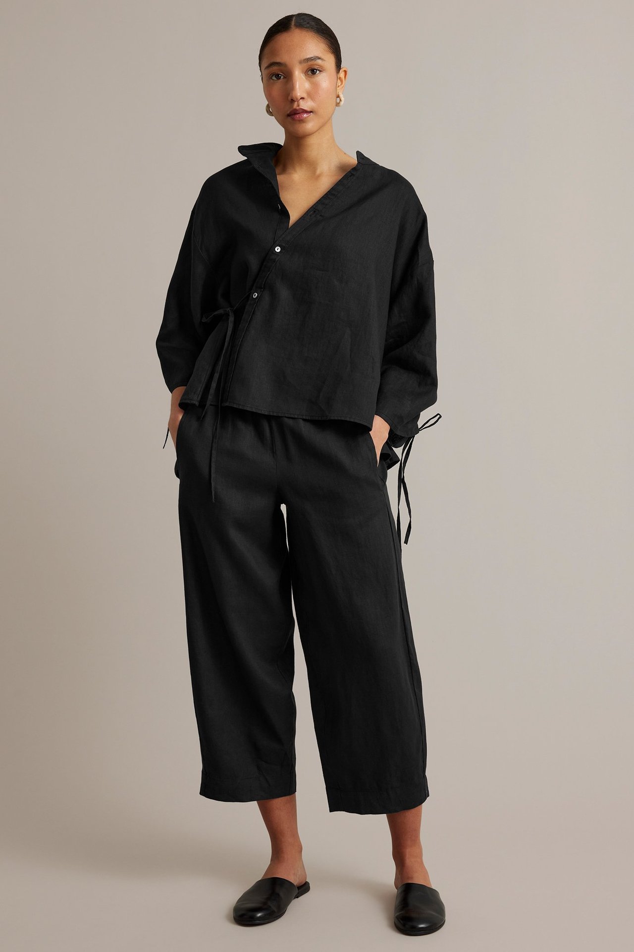 Edestäpäin: Nainen yllään musta pellavapusero vinonapilla, vyötärönauhalla, leveälahkeiset housut ja mustat muulit.