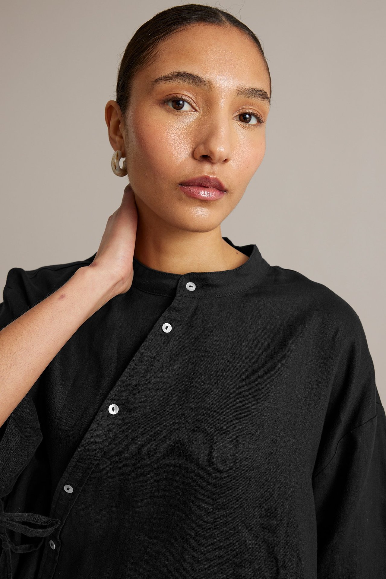 Kvinne i svart linbluse med ståkrage, diagonal knappelukking og lyse bøyleøredobber, forfra.