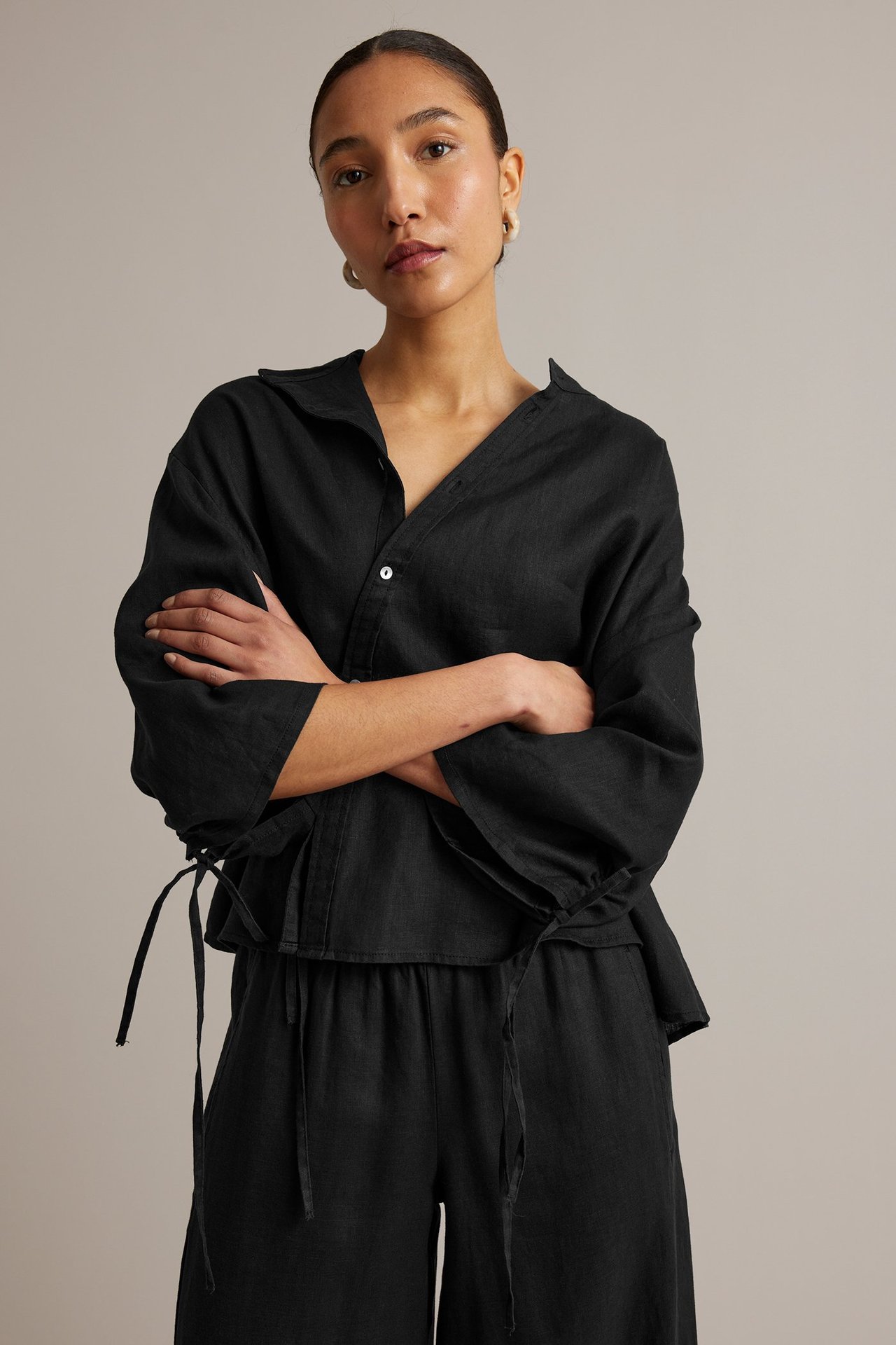 Widok z przodu: Kobieta w czarnej lnianej bluzce z ukośnymi guzikami, wiązaniami na rękawach, w talii, czarne spodnie.