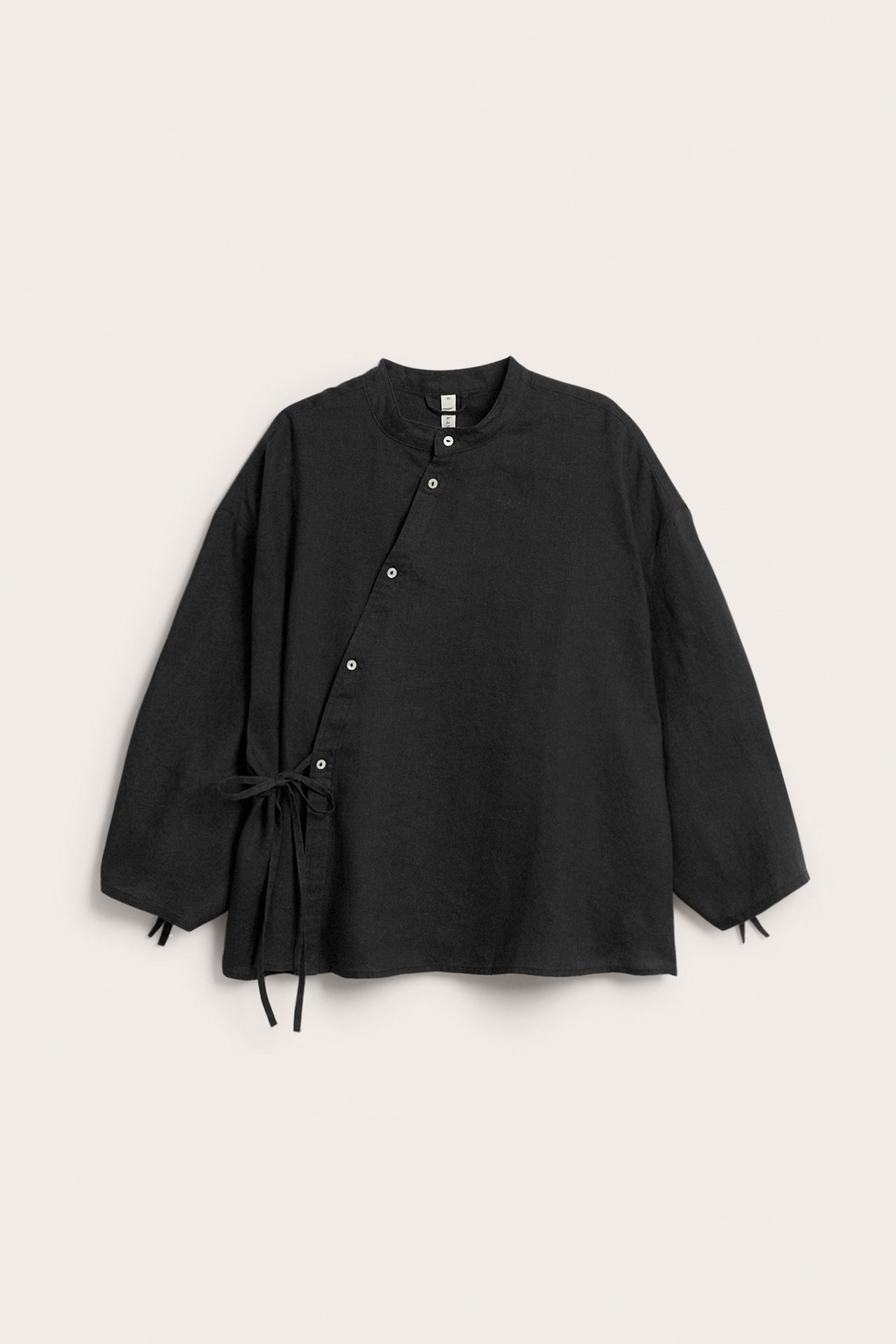 Framifrån: Svart linneblus med ståkrage, asymmetrisk knäppning, knytband i midjan och ärmslut med knytdetaljer.