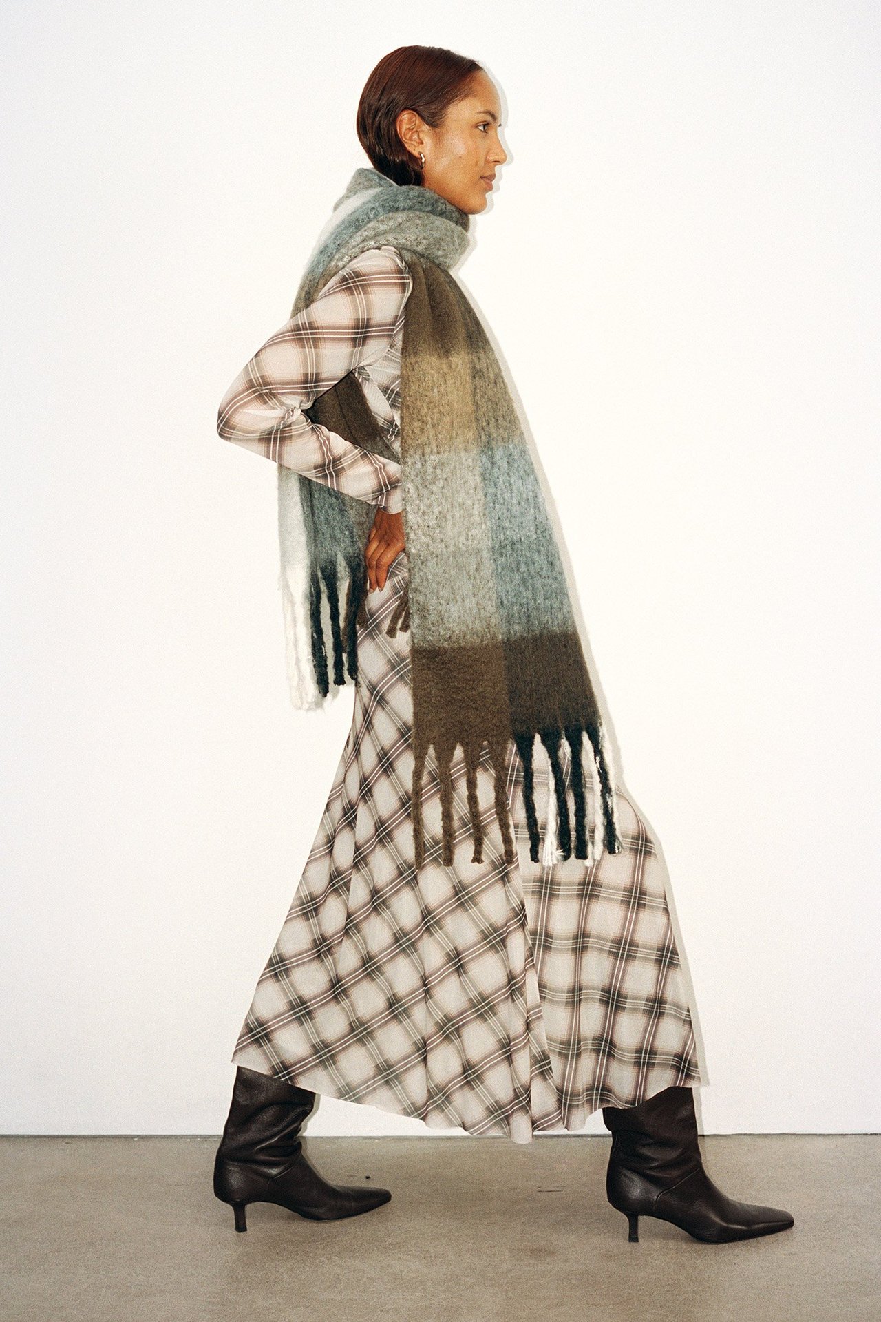Sivukuva: nainen beigessä ruudullisessa epäsymmetrisessä verkkohameessa, läpikuultavassa topissa, värikkäässä huivissa, tummissa saappaissa.