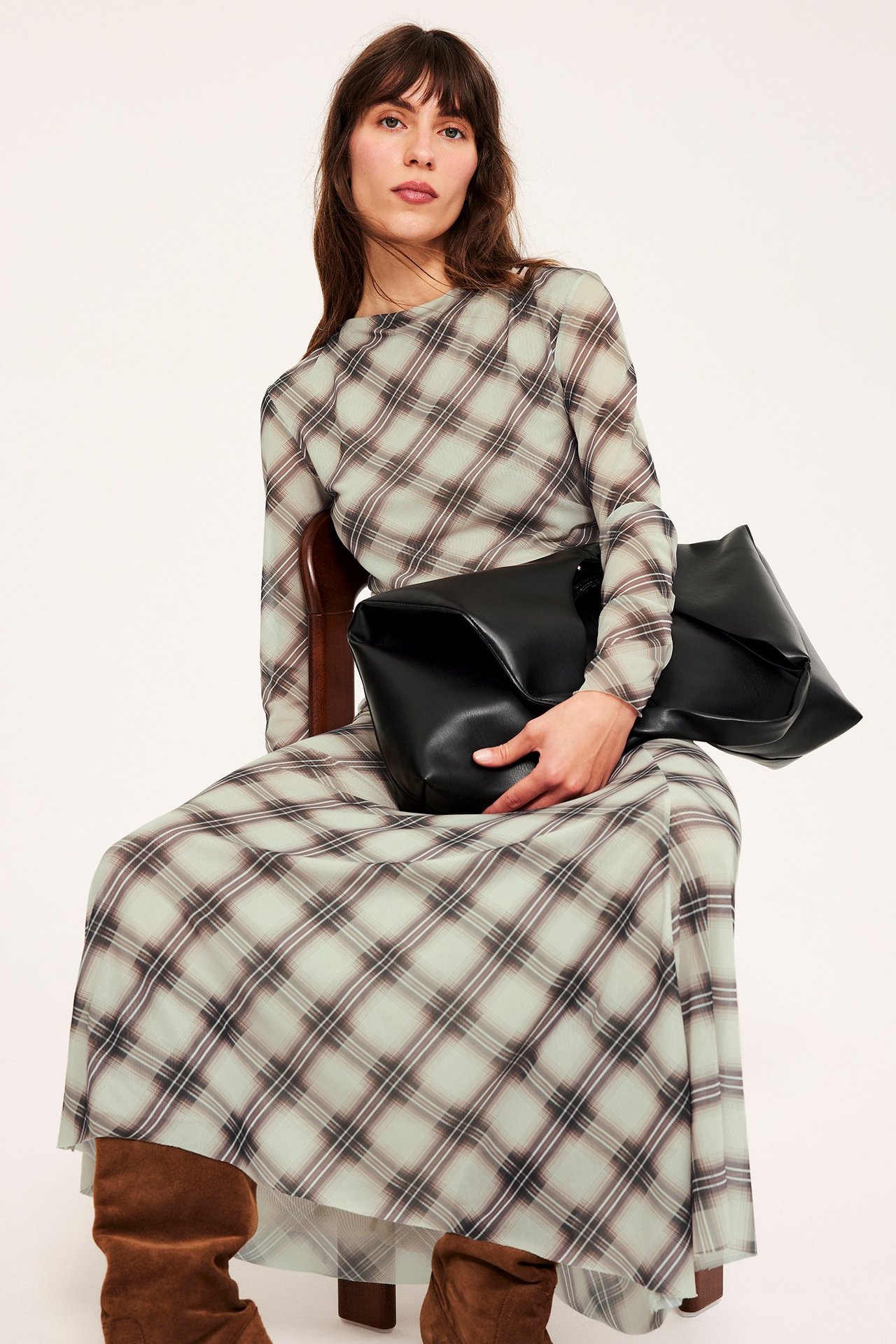 Edestä: Nainen pitkässä beigessä ruudullisessa mekossa, pitää mustaa käsilaukkua, yllään ruskeat saappaat.