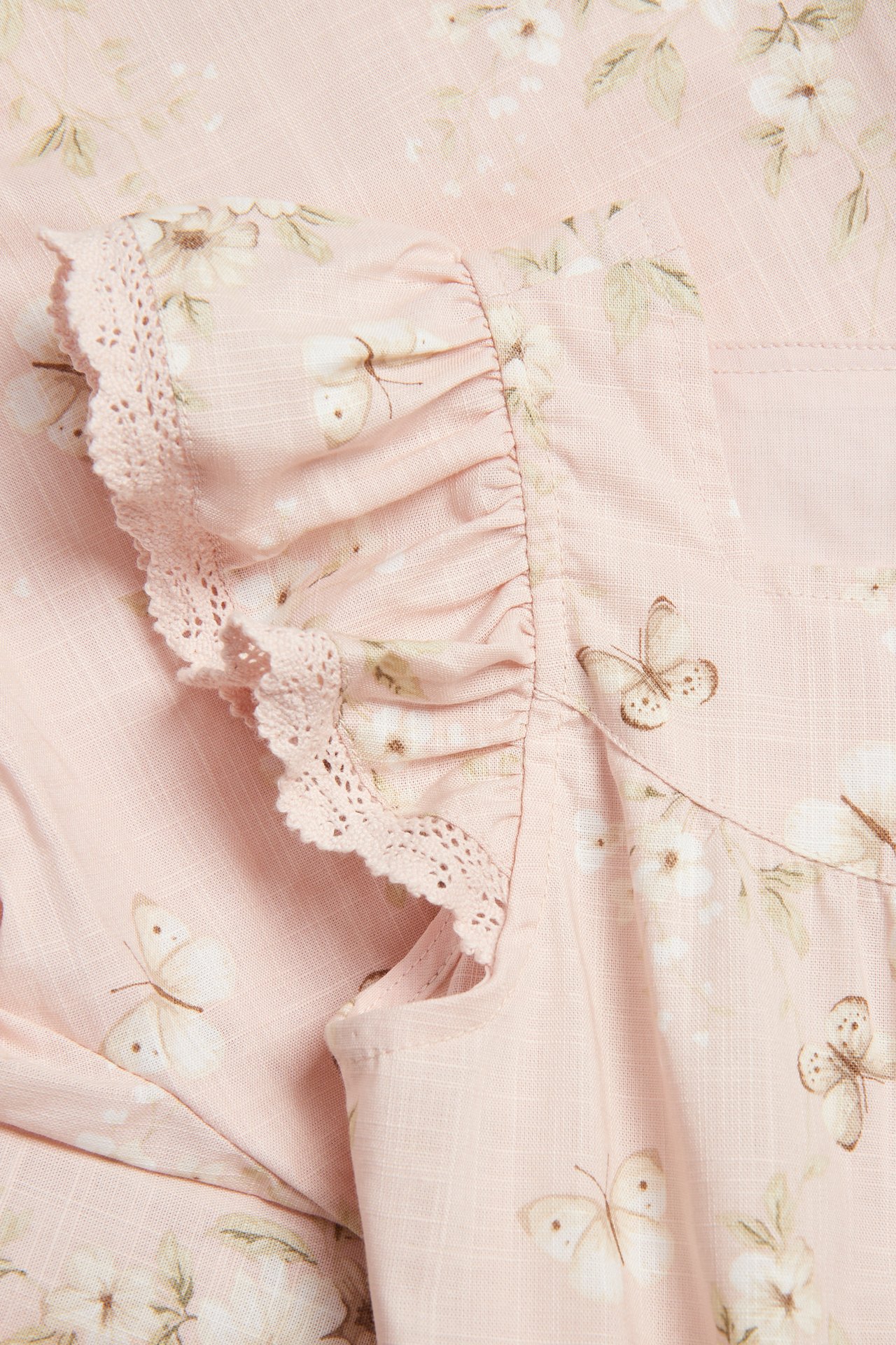Nærbilde av en lyserosa vevd overall med blomster- og sommerfuglmønster, med volangerm med blondekant.