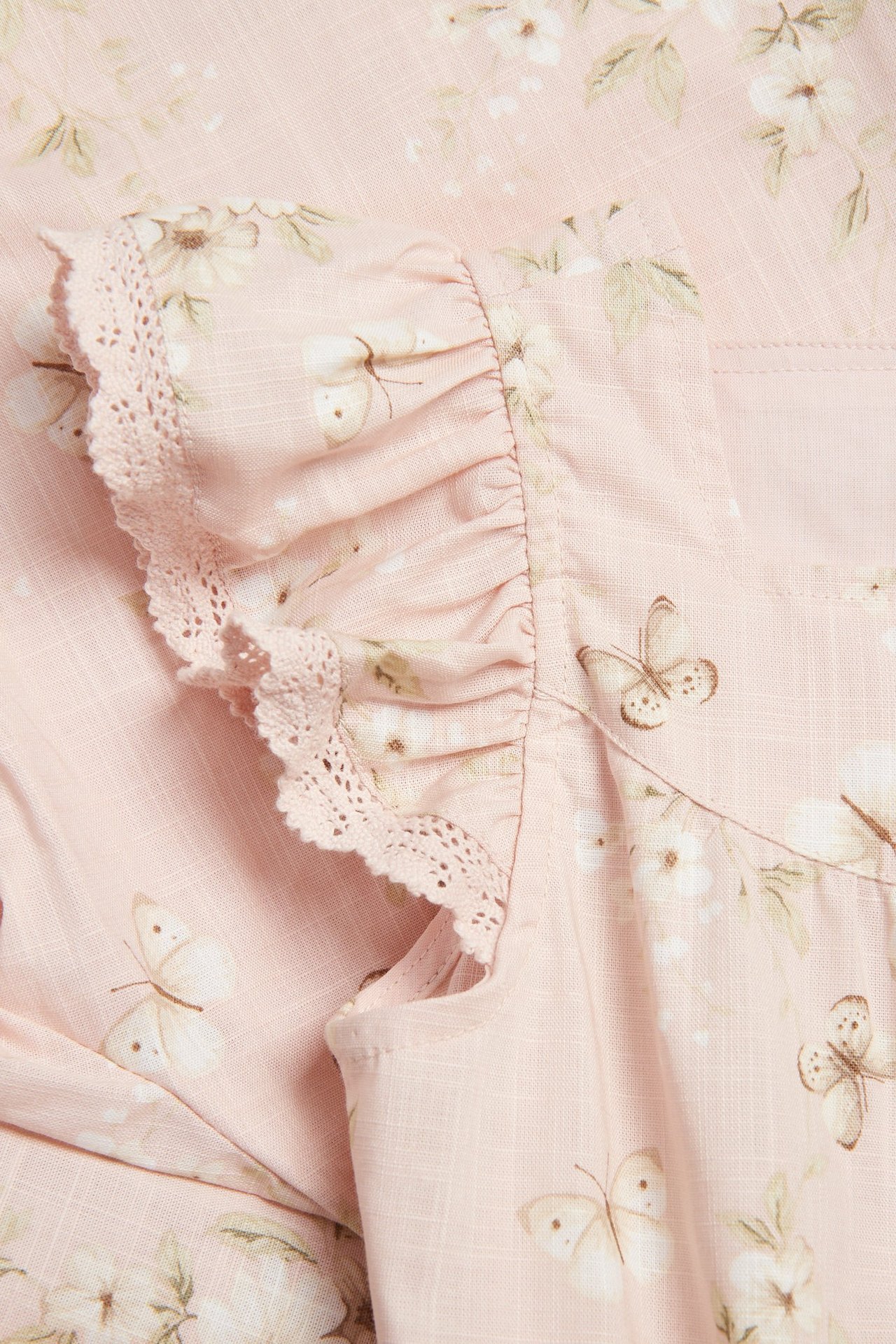 Nærbilde av en lyserosa vevd overall med blomster- og sommerfuglmønster, med volangerm med blondekant.