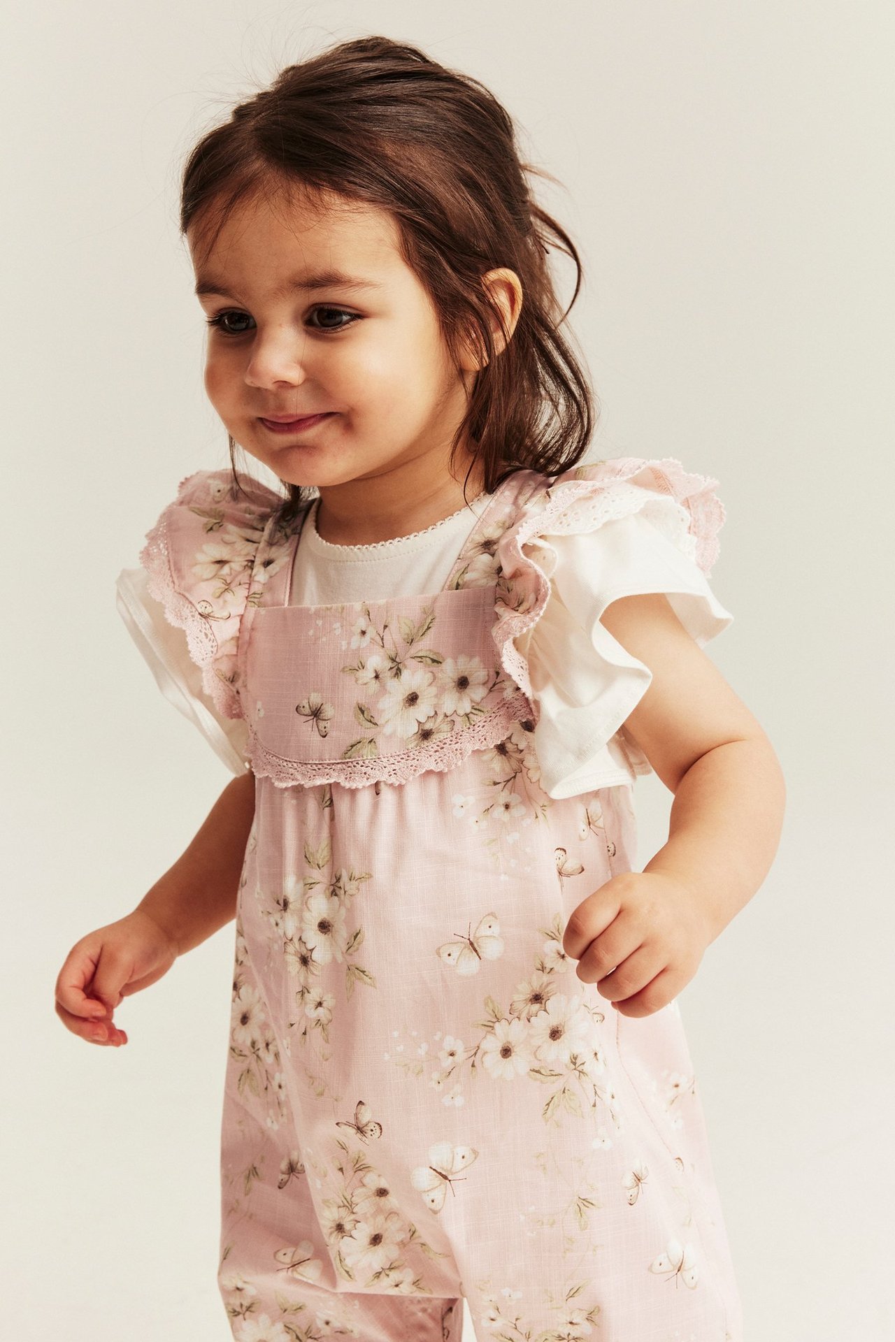 Forfra: Babyjente i en lys rosa blomstret bomullsjumpsuit med volangermer, over en hvit topp.