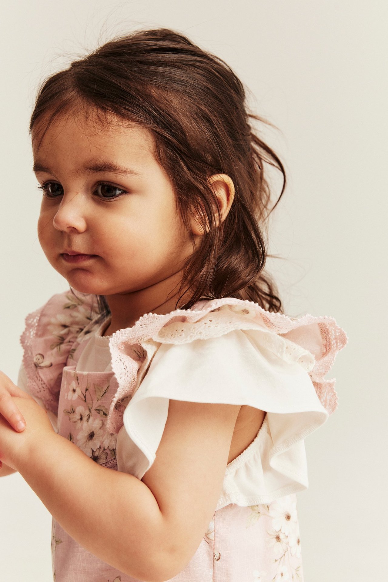 Widok z boku niemowlęcia w jasnoróżowym kombinezonie w kwiaty z falbanami i koronką, na białym topie.