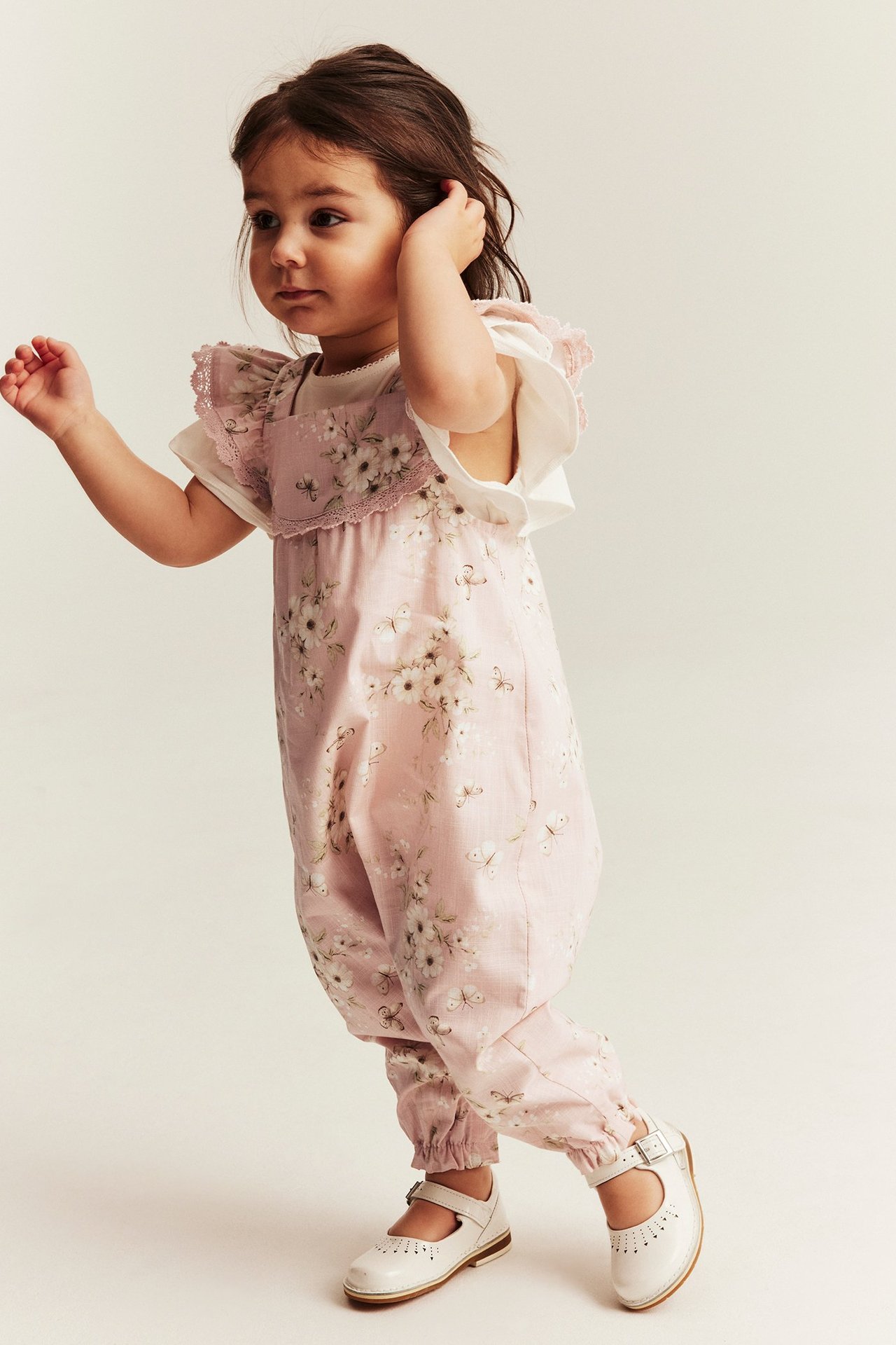 Frontbilde: Barn i lyserosa blomstret bomullsjumpsuit, over hvit t-skjorte og hvite sko.